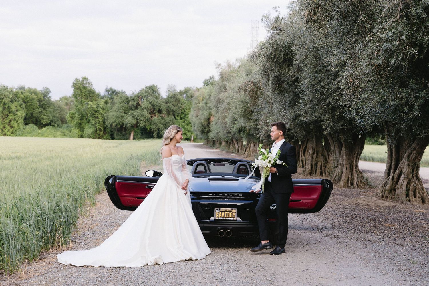 Romantic wedding photo shoot with couple and luxury car on a tree-lined path.