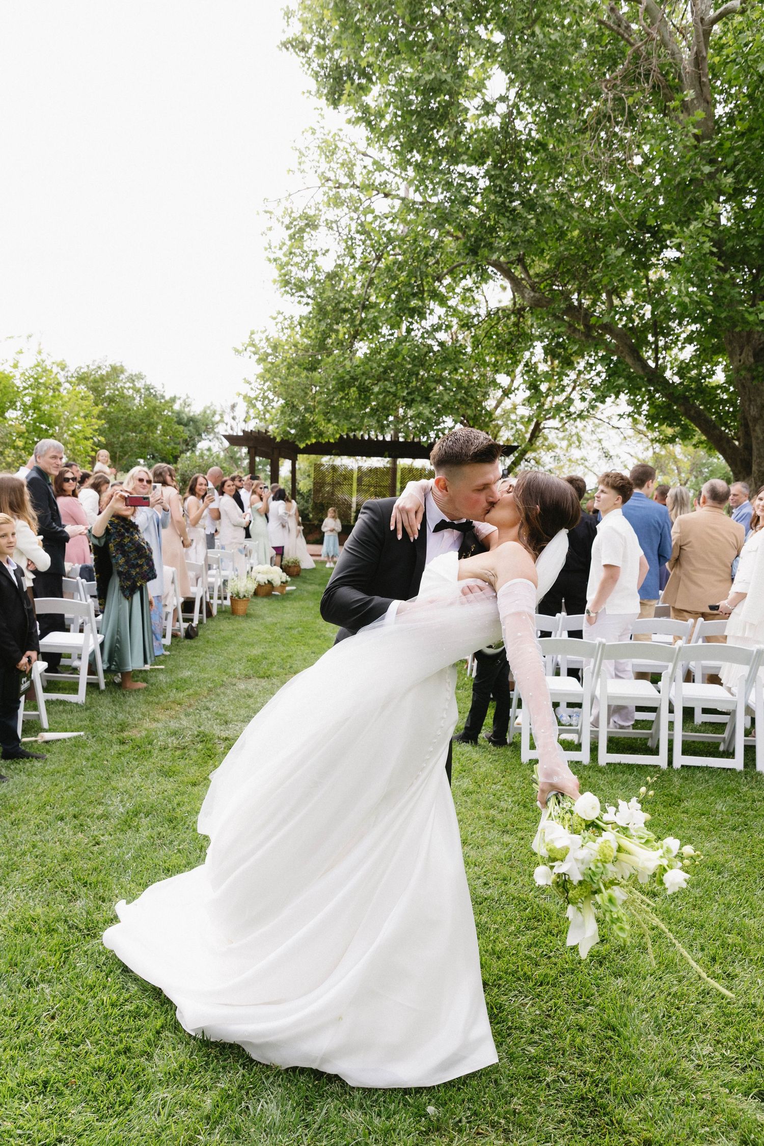 Newlyweds dancing together on a green lawn with white chairs and wedding guests in the background.