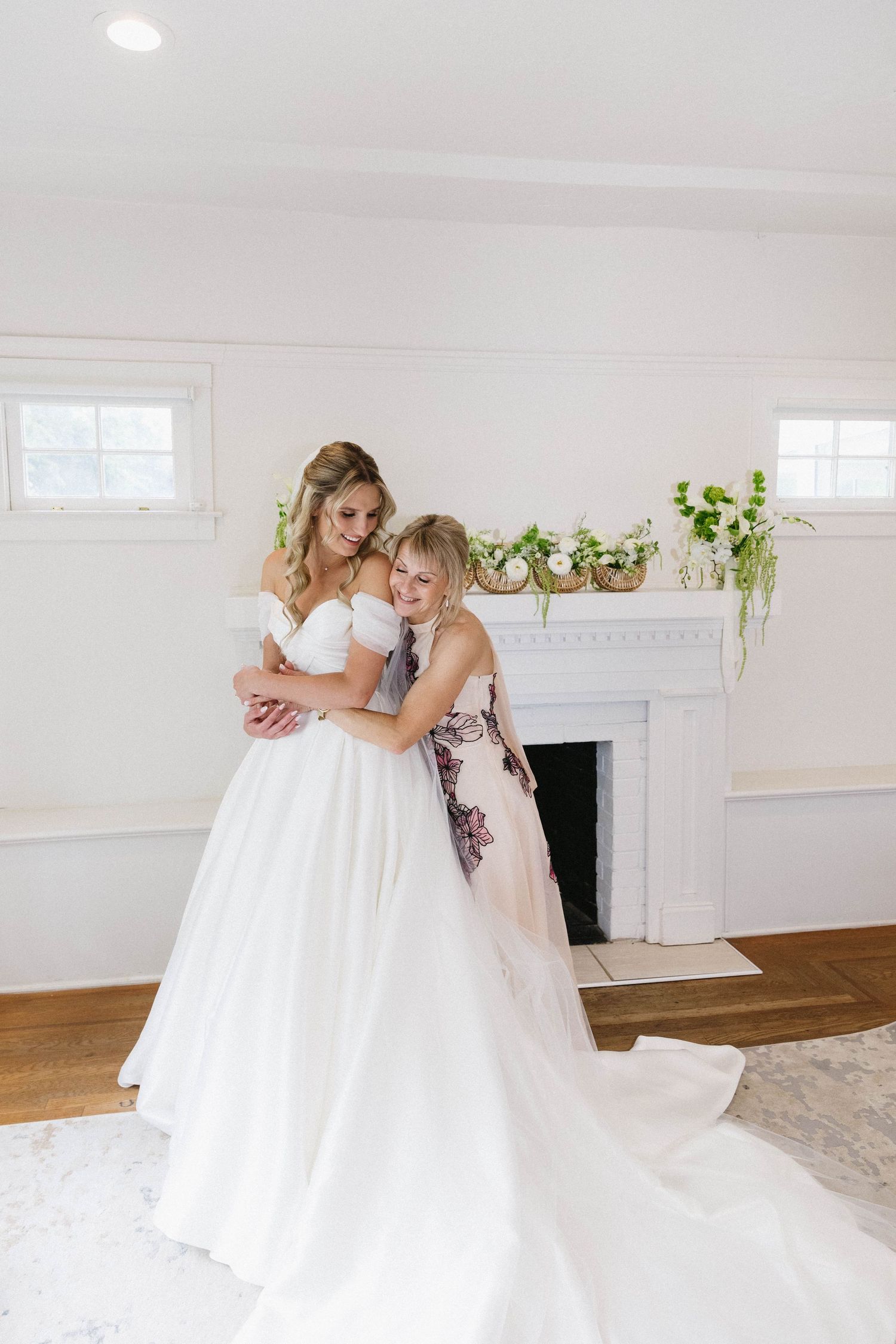 Tender embrace during wedding dress fitting in bright room with white fireplace.