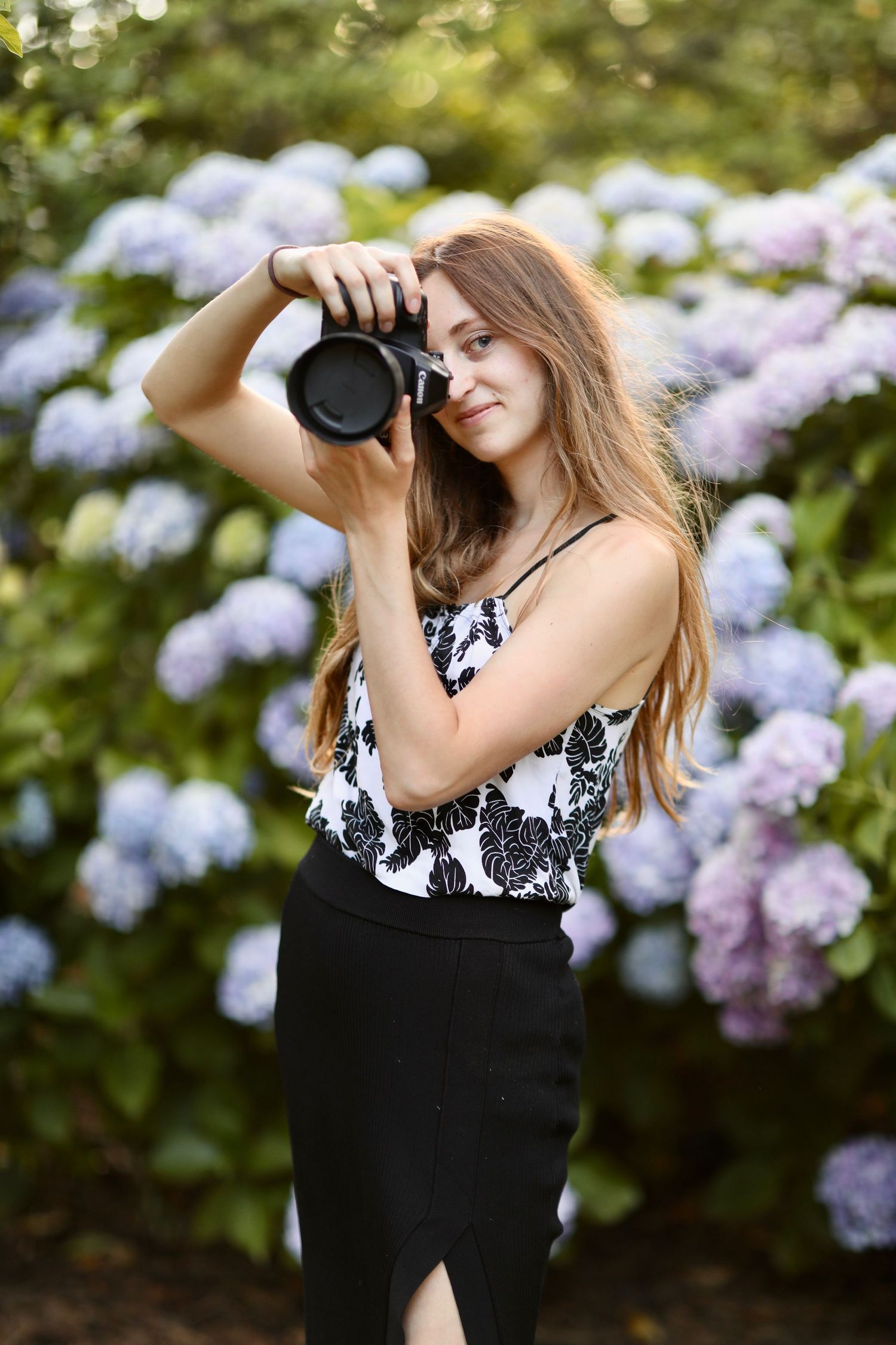 Photographer with long hair takes pictures in front of blooming purple hydrangea bushes wearing black and white outfit.
