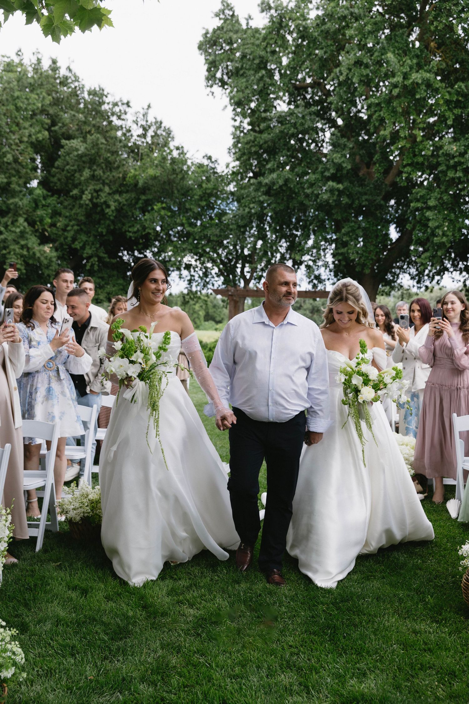 Wedding party walking down grassy aisle after ceremony with guests seated in white chairs.