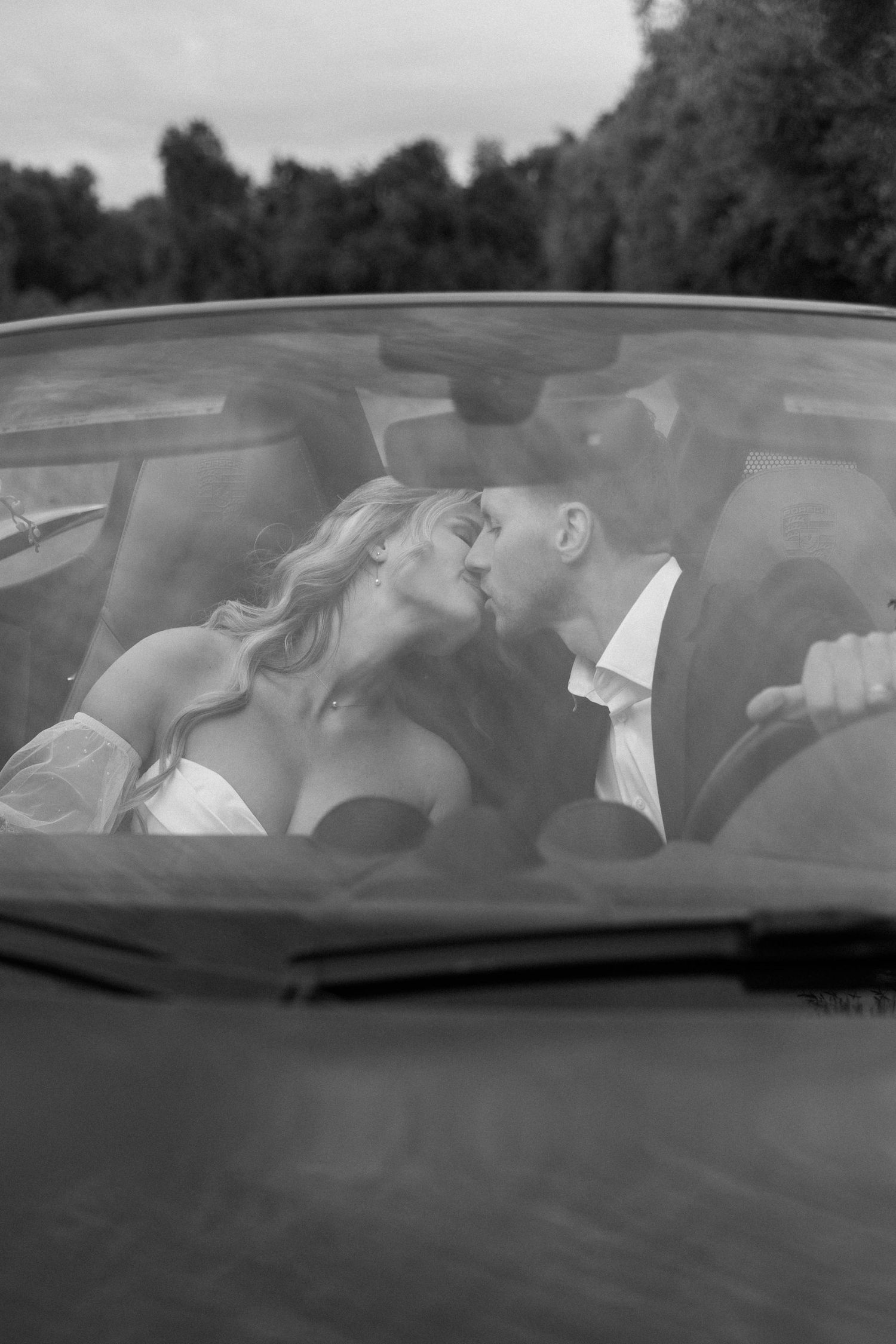 Black and white photo of newlyweds sharing an intimate moment through a car window.