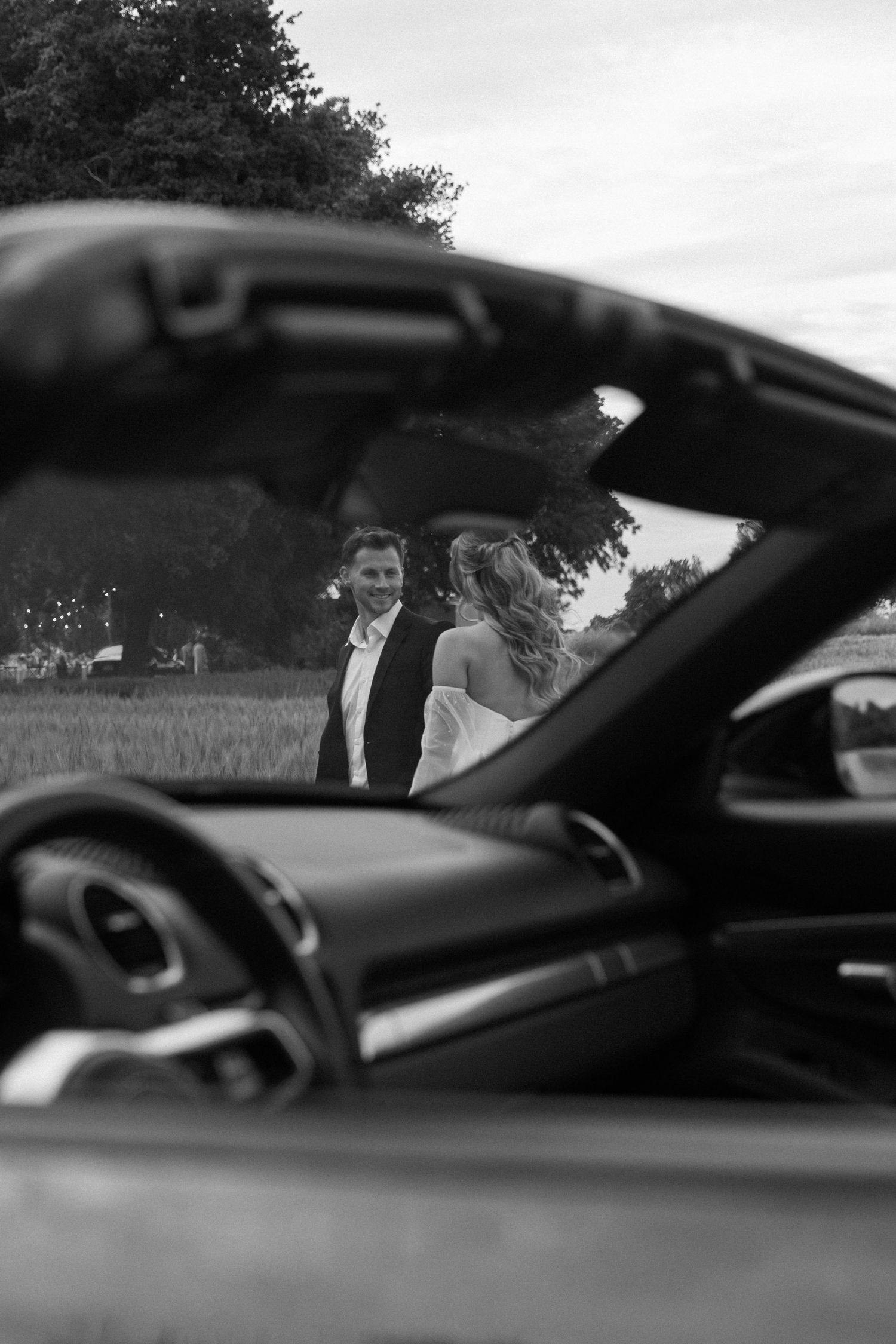Black and white photo through car window showing couple by convertible.