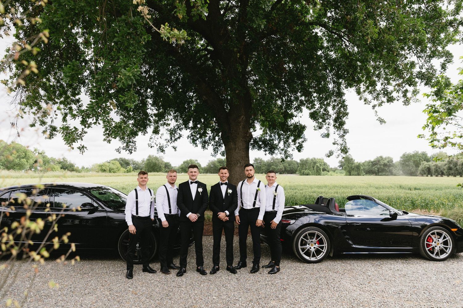 Wedding party posing with luxury cars in an outdoor setting with trees in the background.