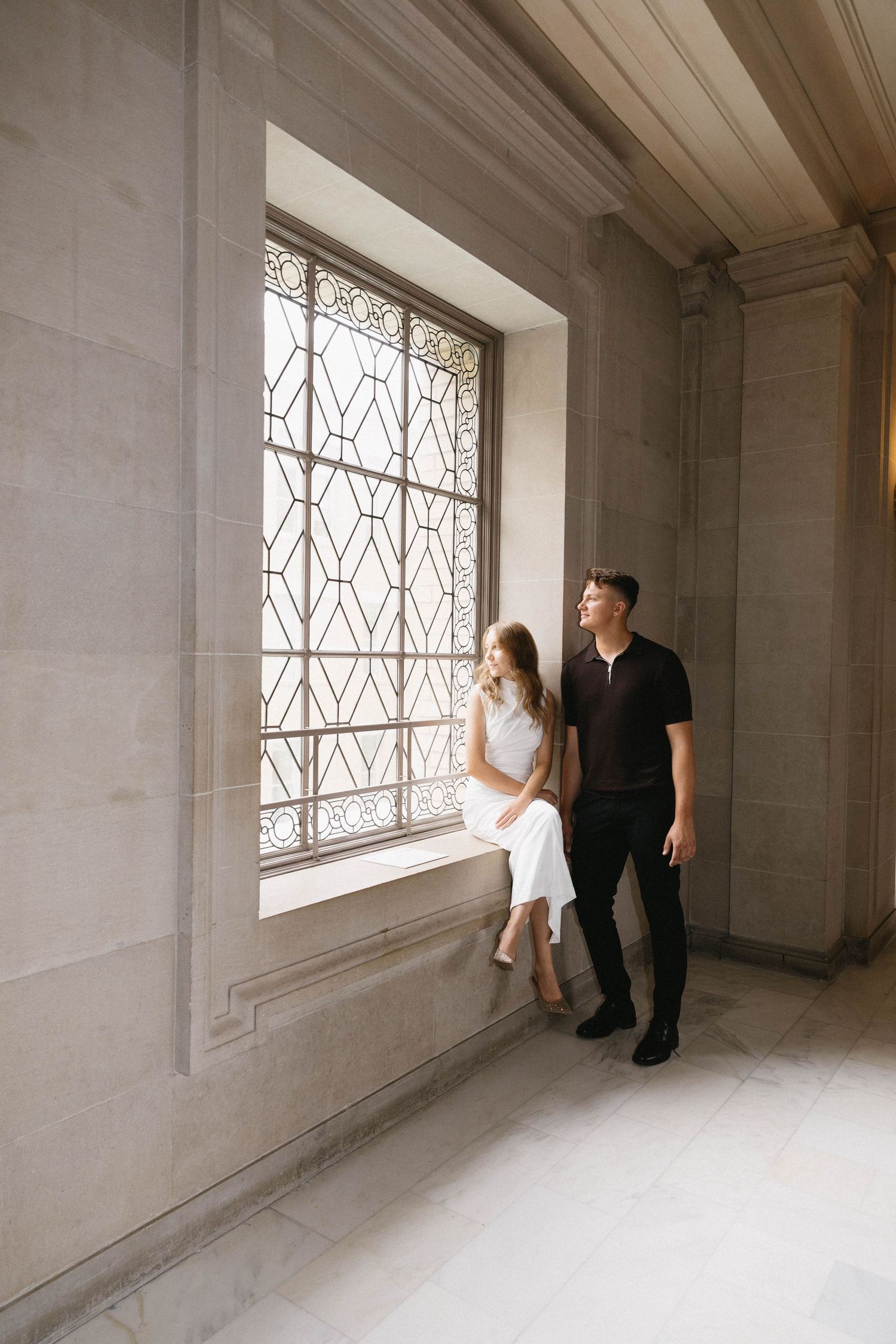 A couple stands in a minimalist white room with architectural windows creating a dramatic light-filled composition.