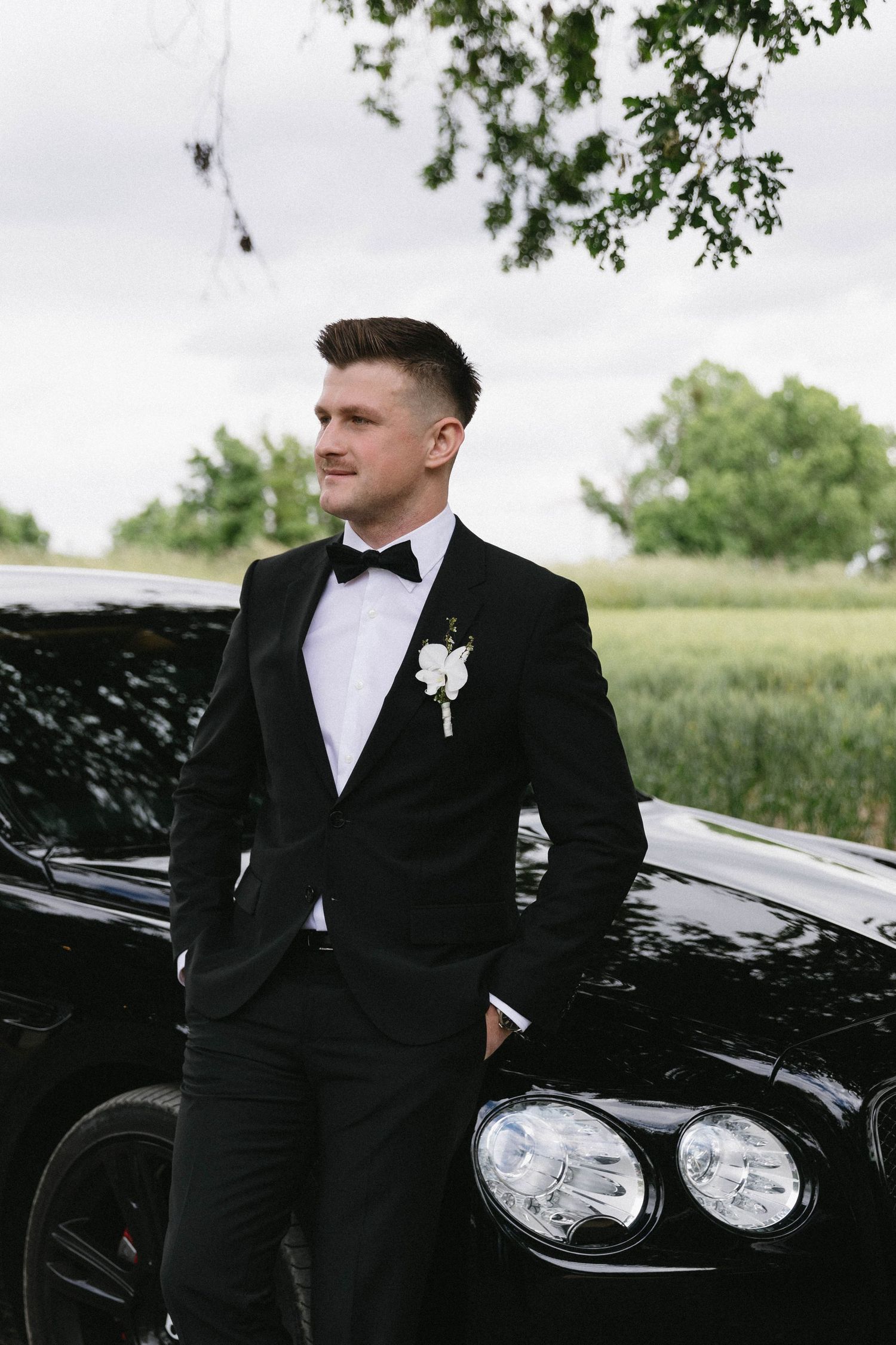 Stylish groom in black tuxedo poses against a sleek black luxury vehicle outdoors.