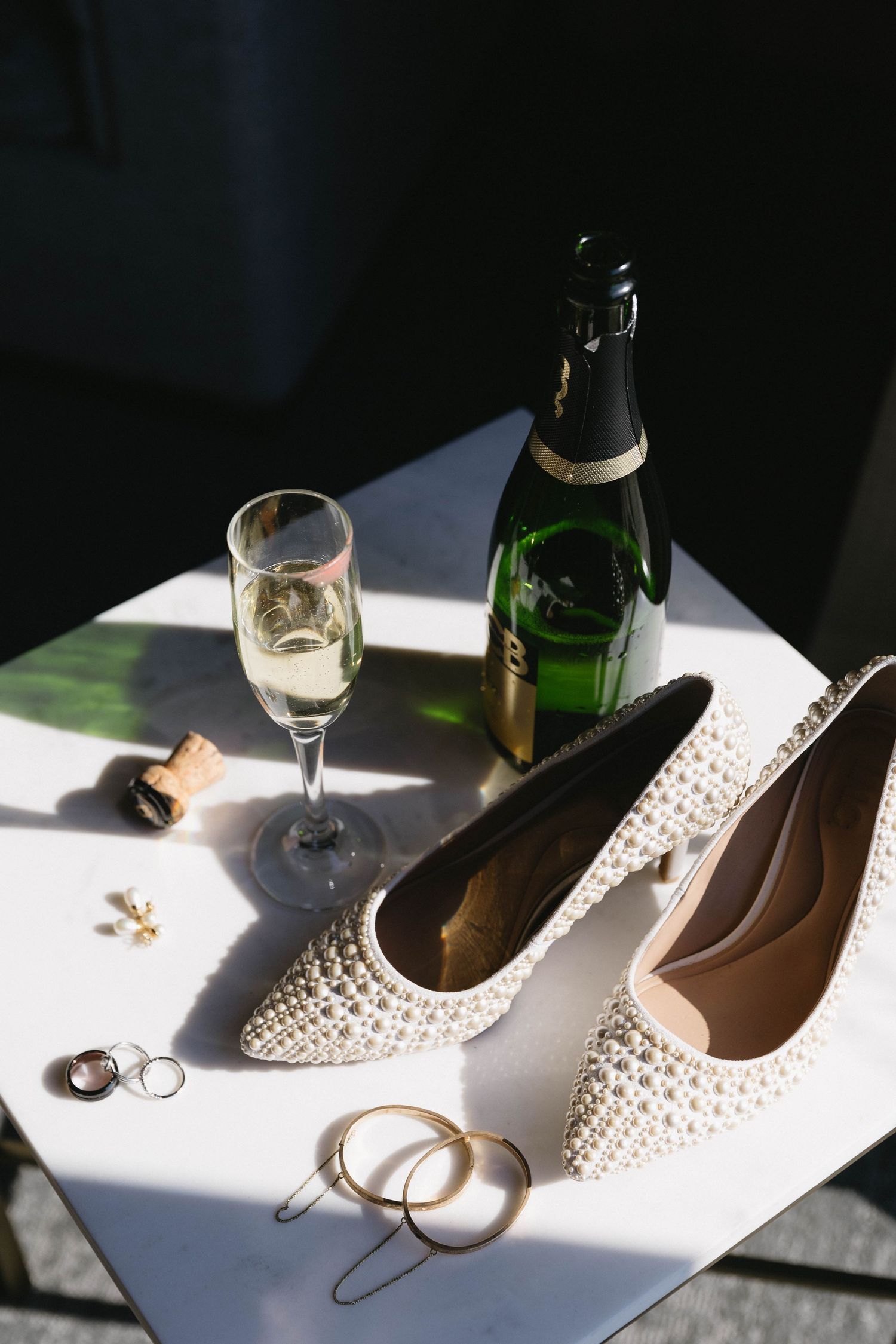 Elegant white pearl-studded bridal shoes and champagne glasses arranged in dramatic natural lighting.