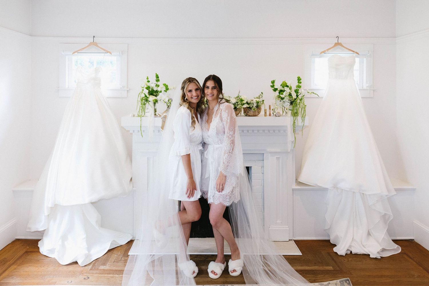 Two wedding dresses displayed on hangers in a bright white room with greenery decorations.