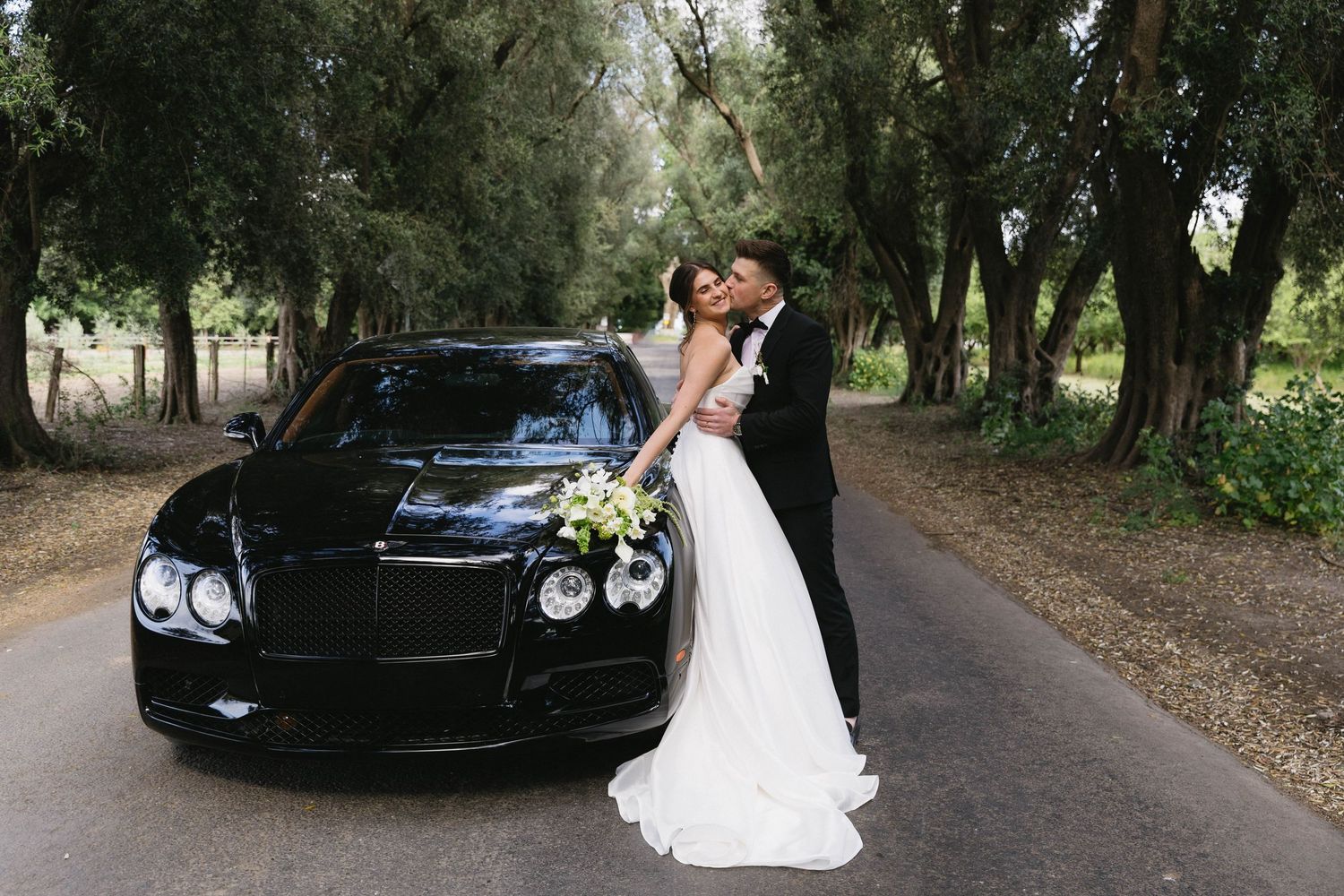 A couple in wedding attire embraces next to a black luxury car on a tree-lined road.
