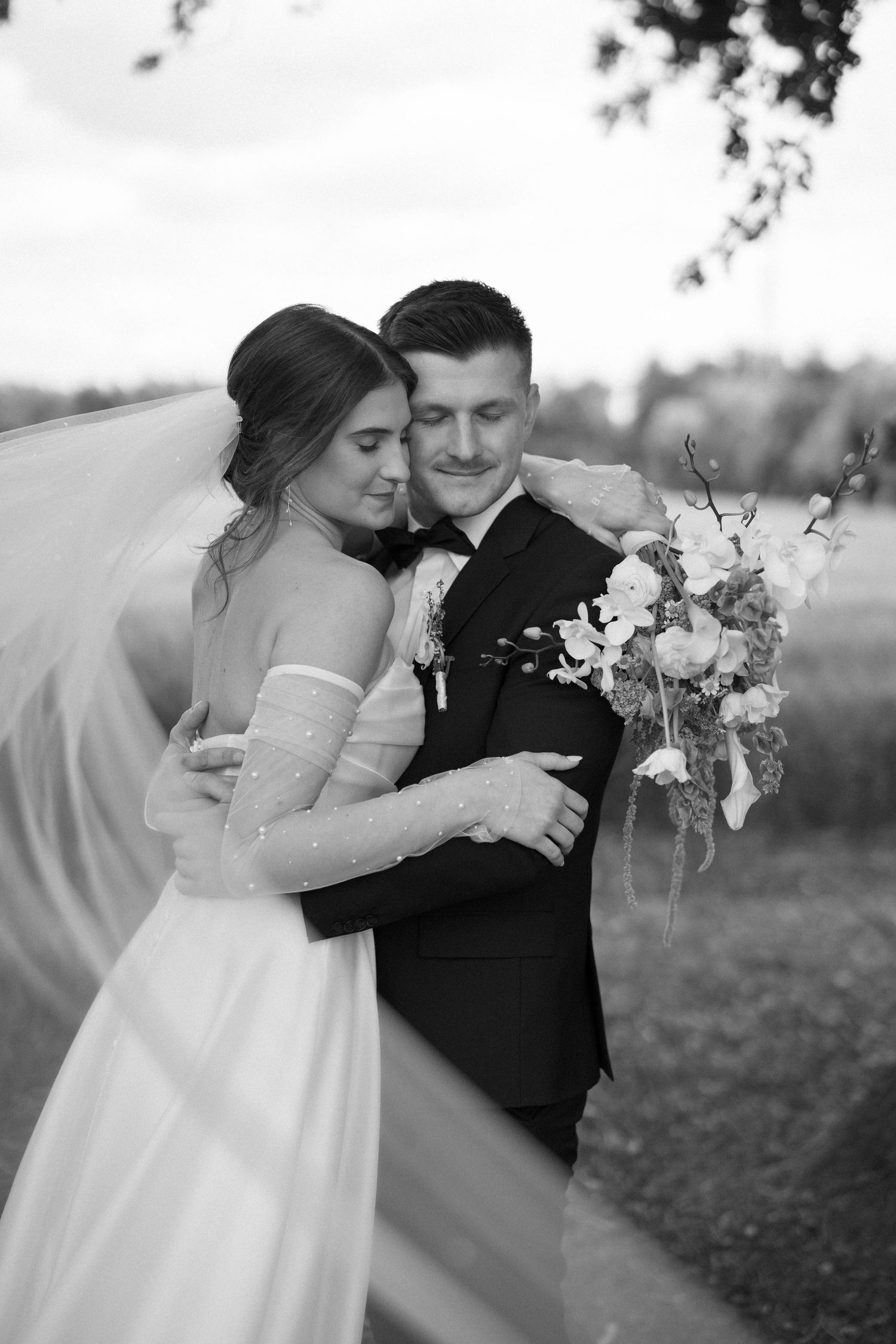 A romantic black and white wedding portrait of a couple embracing outdoors with the bride holding a floral bouquet.