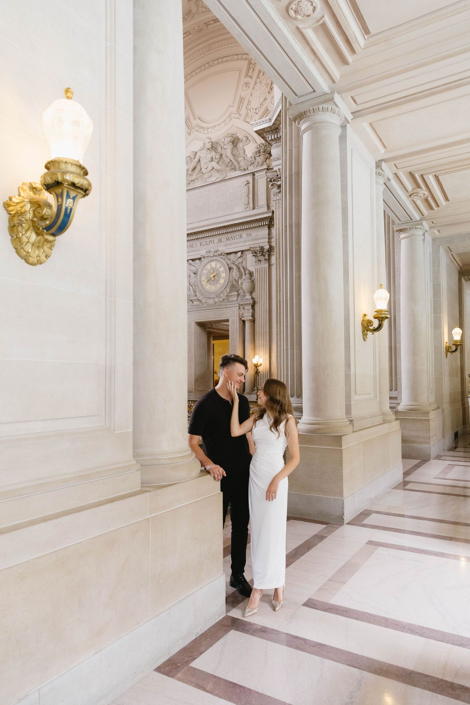 A couple walks through a luxurious white marble hallway with ornate gold wall sconces.