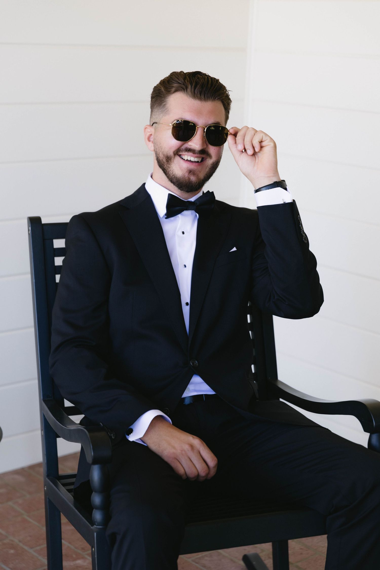 A groom in a classic black tuxedo and sunglasses poses casually in a wooden chair.