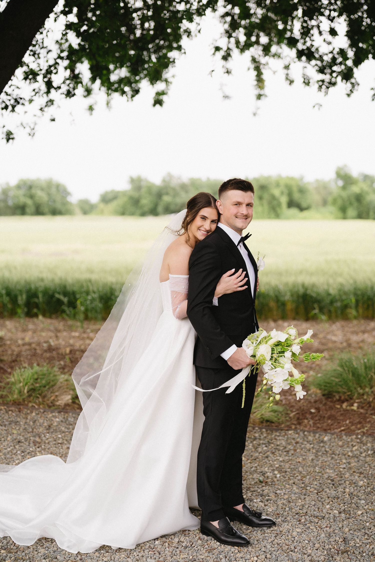 Full-length wedding portrait of couple standing on a gravel path with trees and field in background.