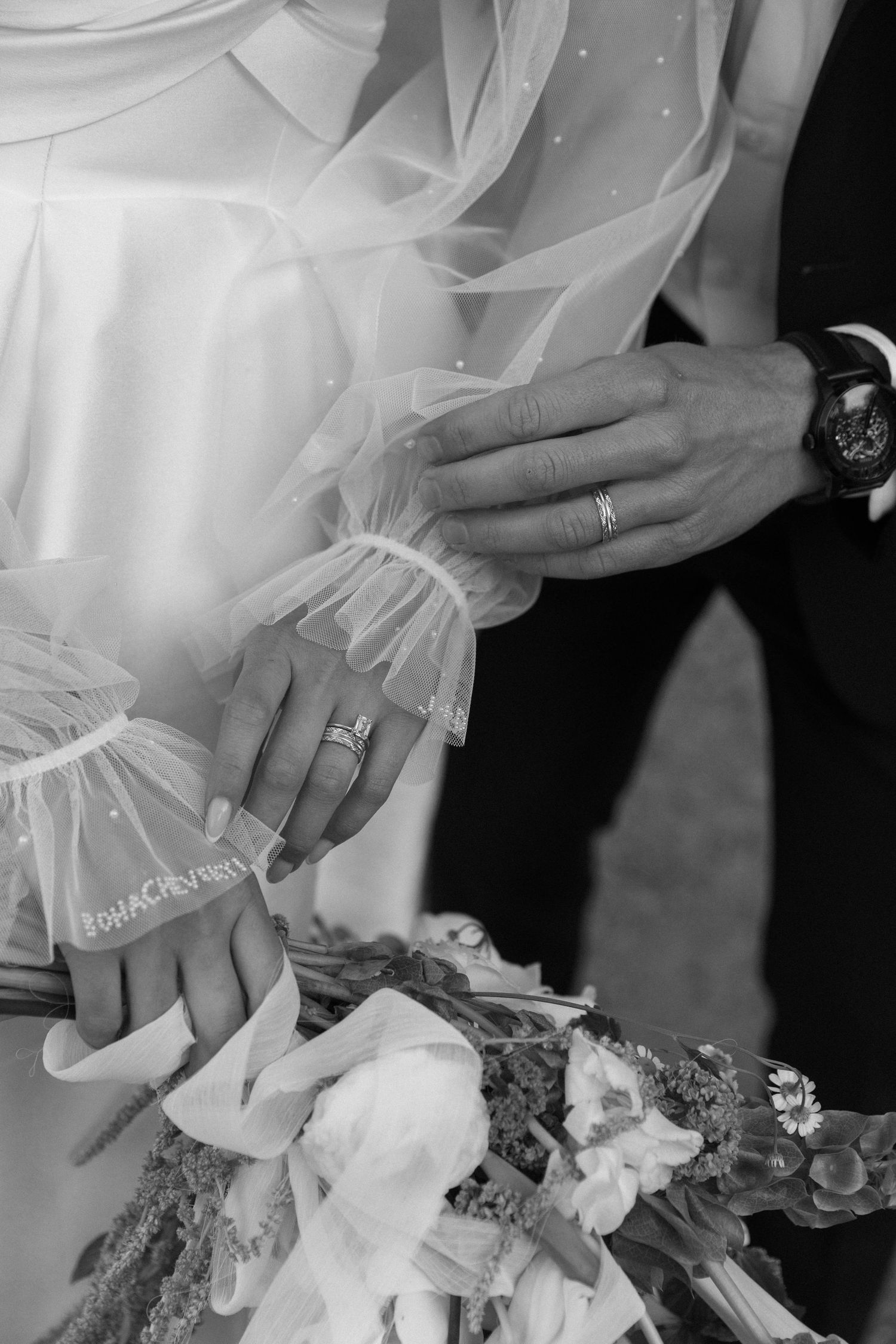 Close-up black and white photo of wedding rings and hands against white dress fabric.