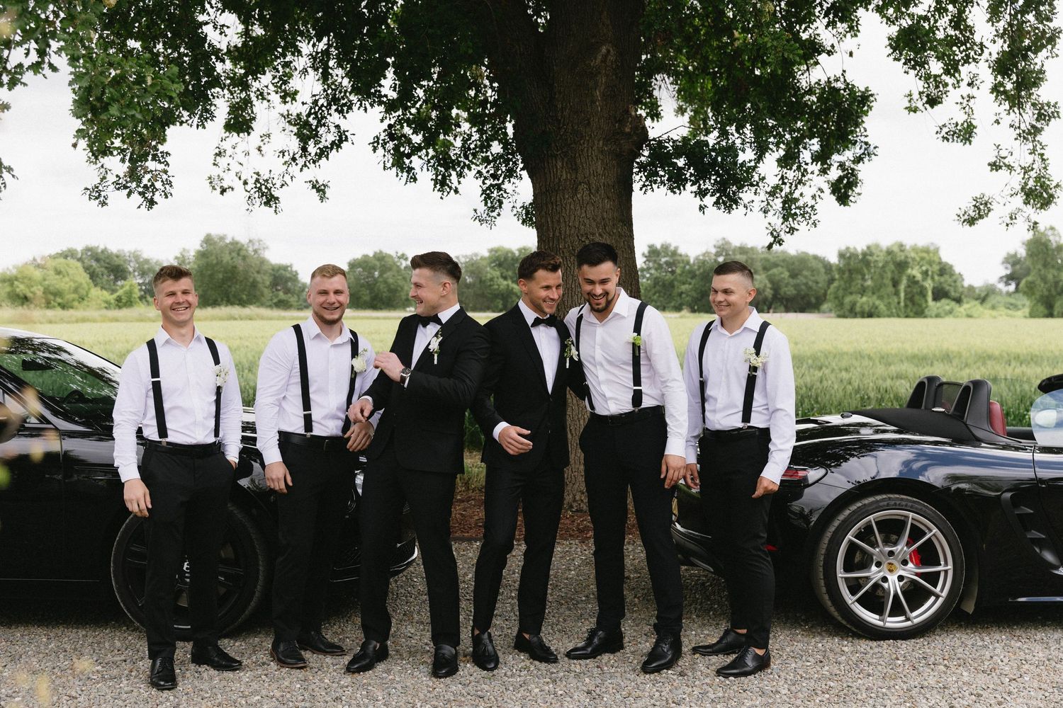 Groomsmen in black and white attire pose together near luxury vehicles outdoors.