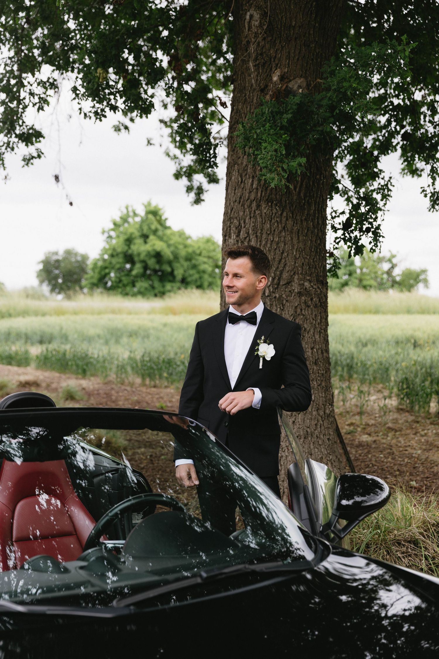 Groom poses with black luxury convertible sports car in outdoor wedding photos.