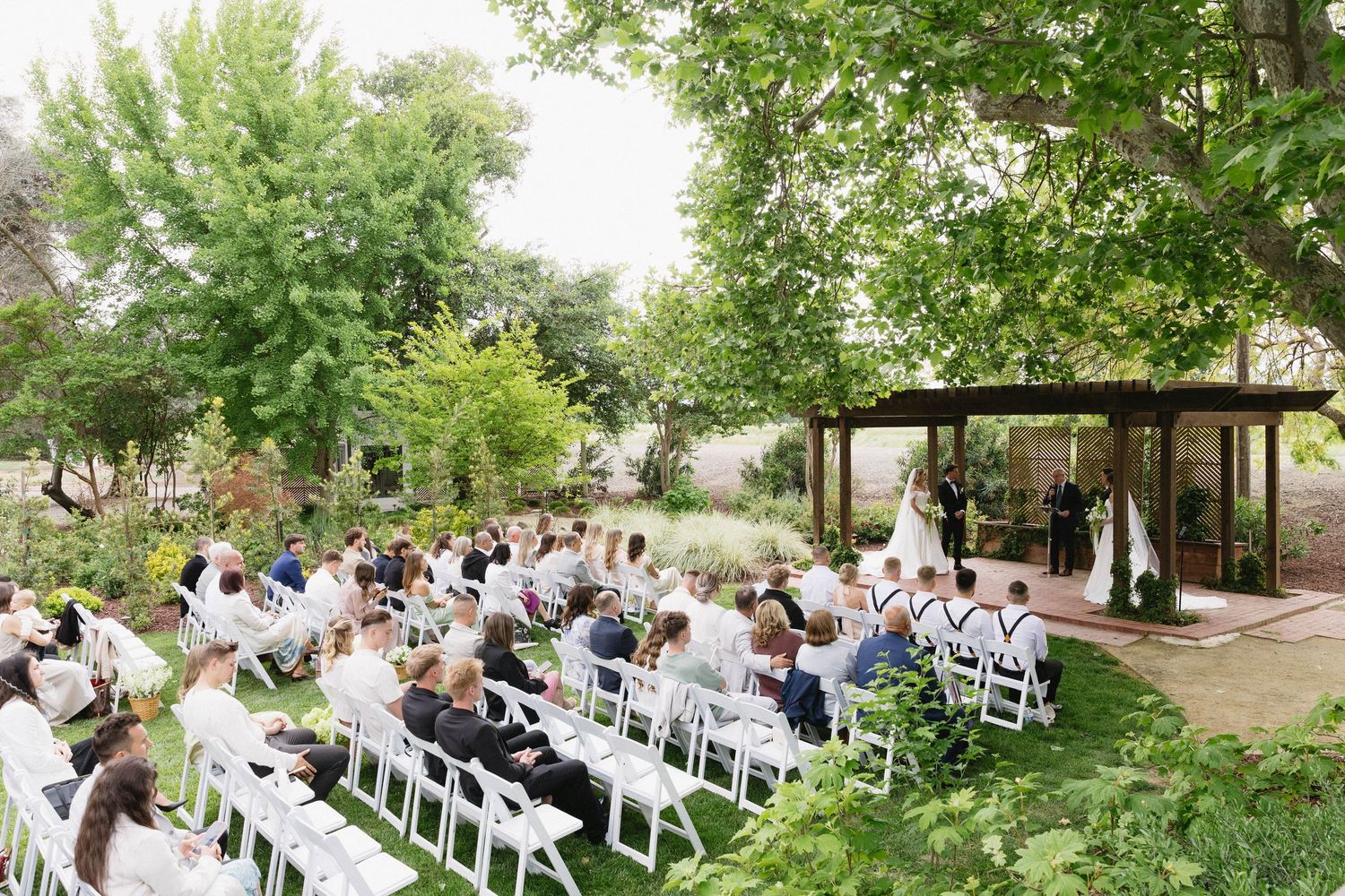 Garden wedding ceremony setup with rows of white chairs leading to wooden pergola.