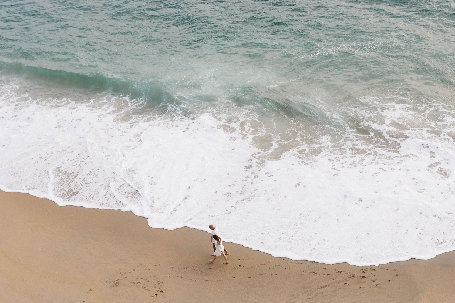 Aerial view of turquoise ocean waves crashing onto a sandy beach with white foam patterns along the shoreline.