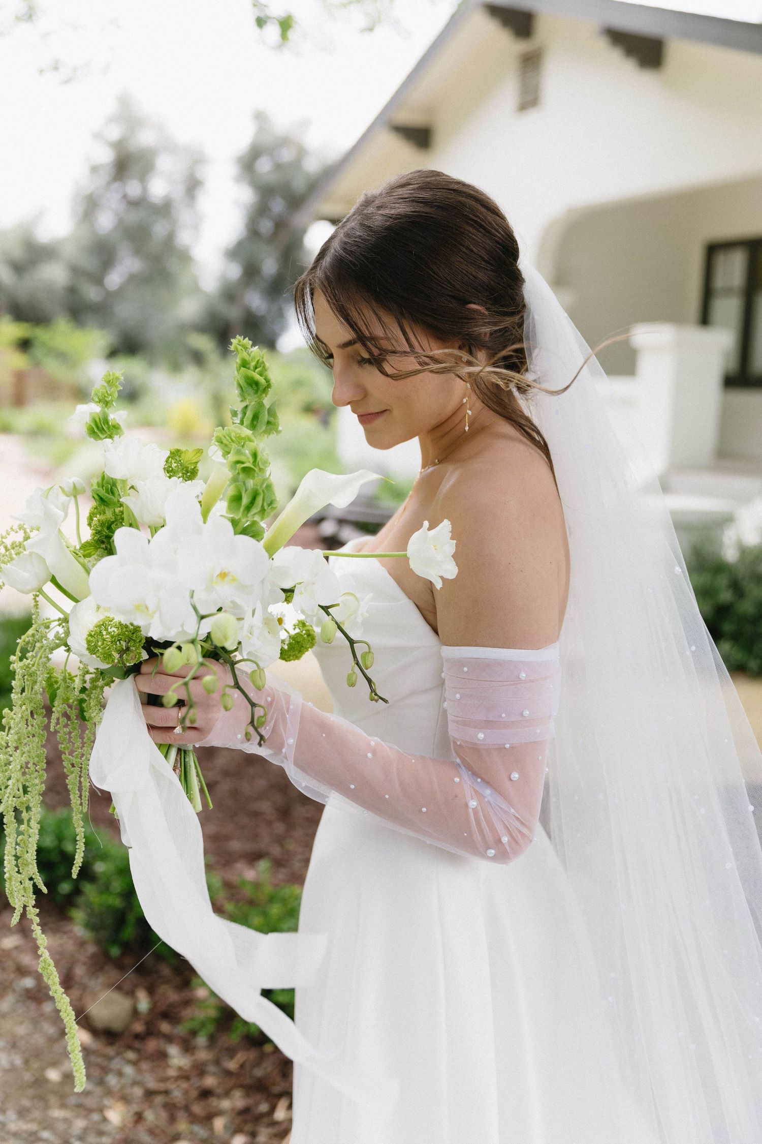 Bride in white lace gown with sheer sleeves holding delicate floral bouquet.