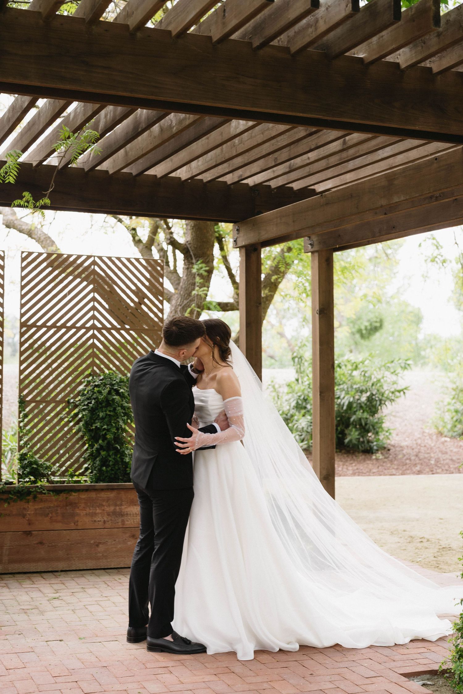 Romantic wedding moment under a wooden pergola with brick paving.
