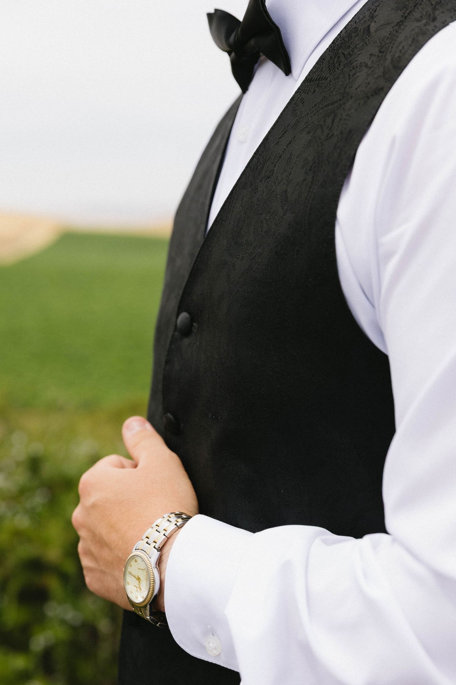 Close-up of formal wedding attire showing black vest, bowtie, and gold watch.
