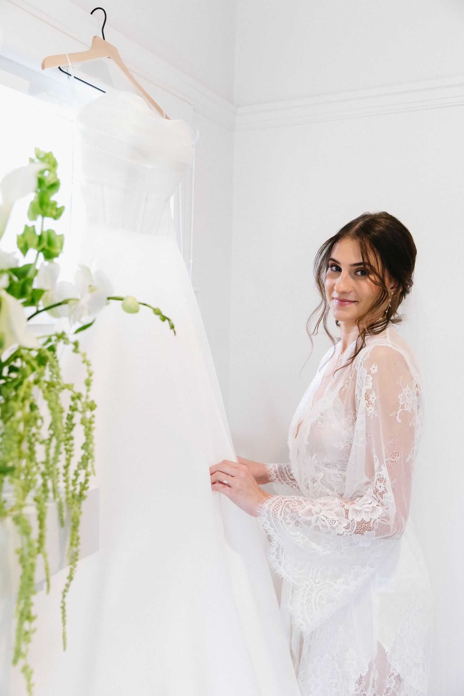 Elegant white lace bridal robe shown against wedding dress in bright room with hanging greenery.