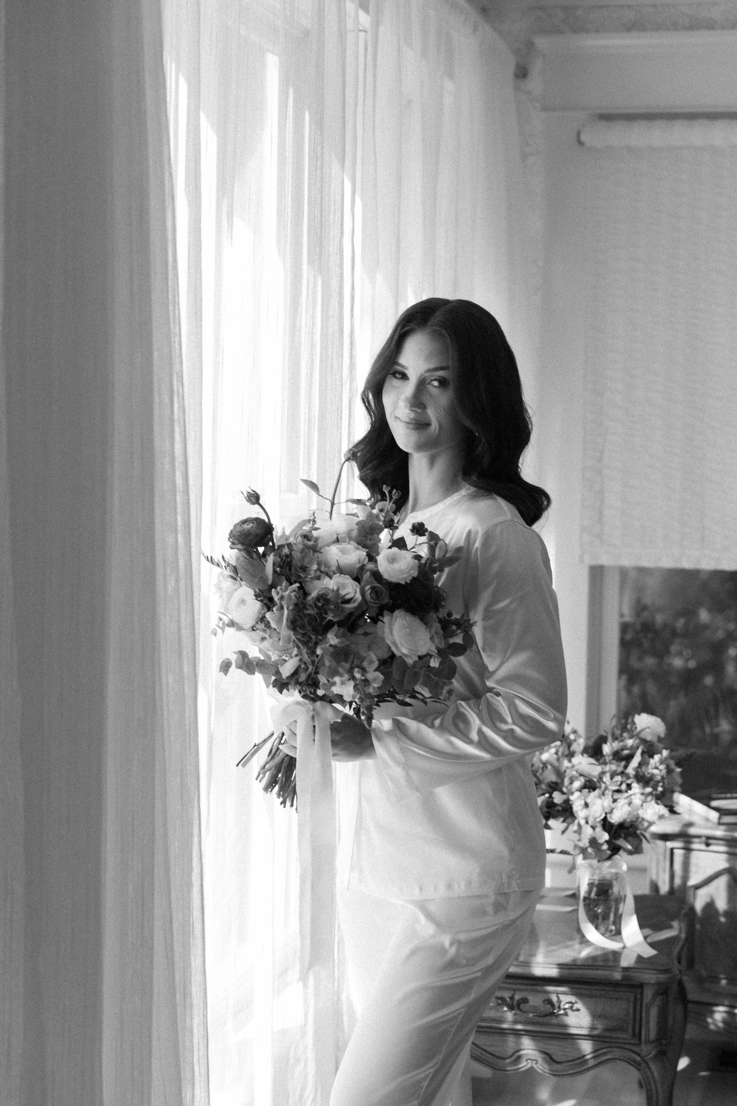 Black and white photo of wedding preparation with floral bouquets in a light-filled room.