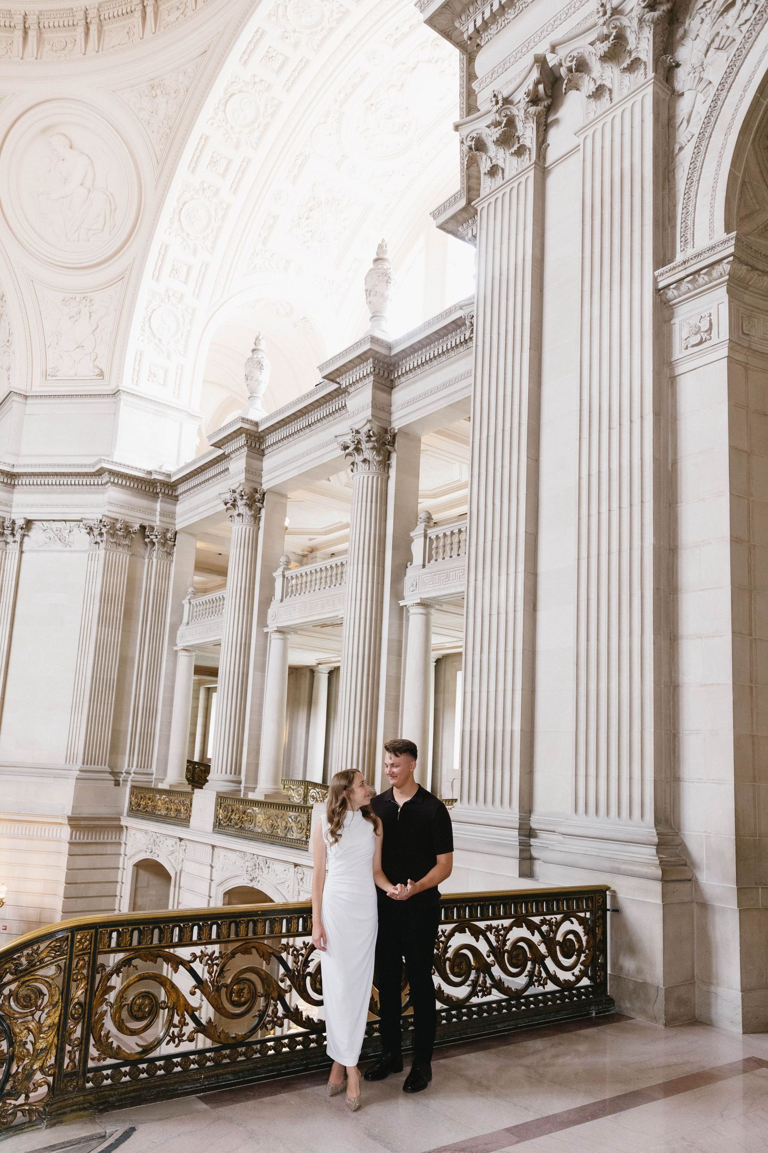 Two people stand together in a grand hallway with classical white columns and decorative gold railings.