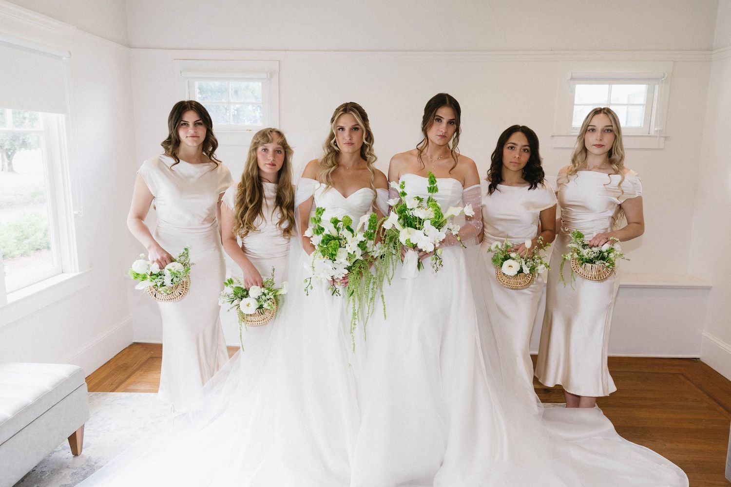 Wedding party in white dresses holding bouquets pose in bright room with hardwood floors.