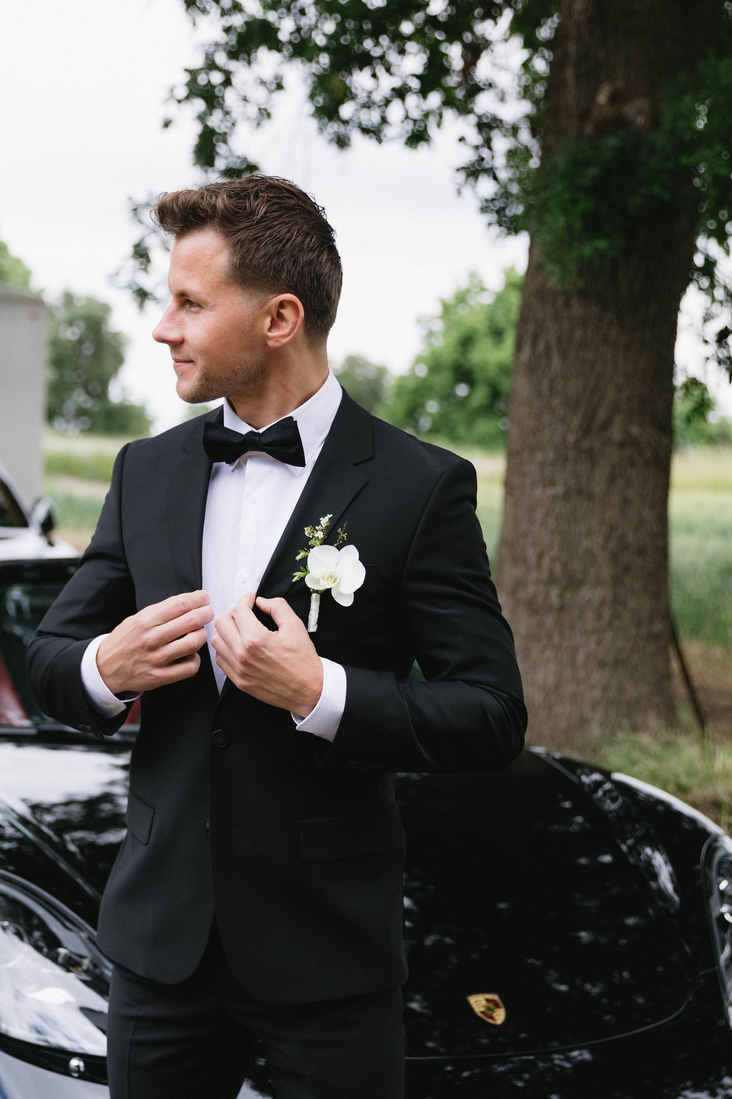 Groom in classic black tuxedo adjusting his tie next to a luxury black car.