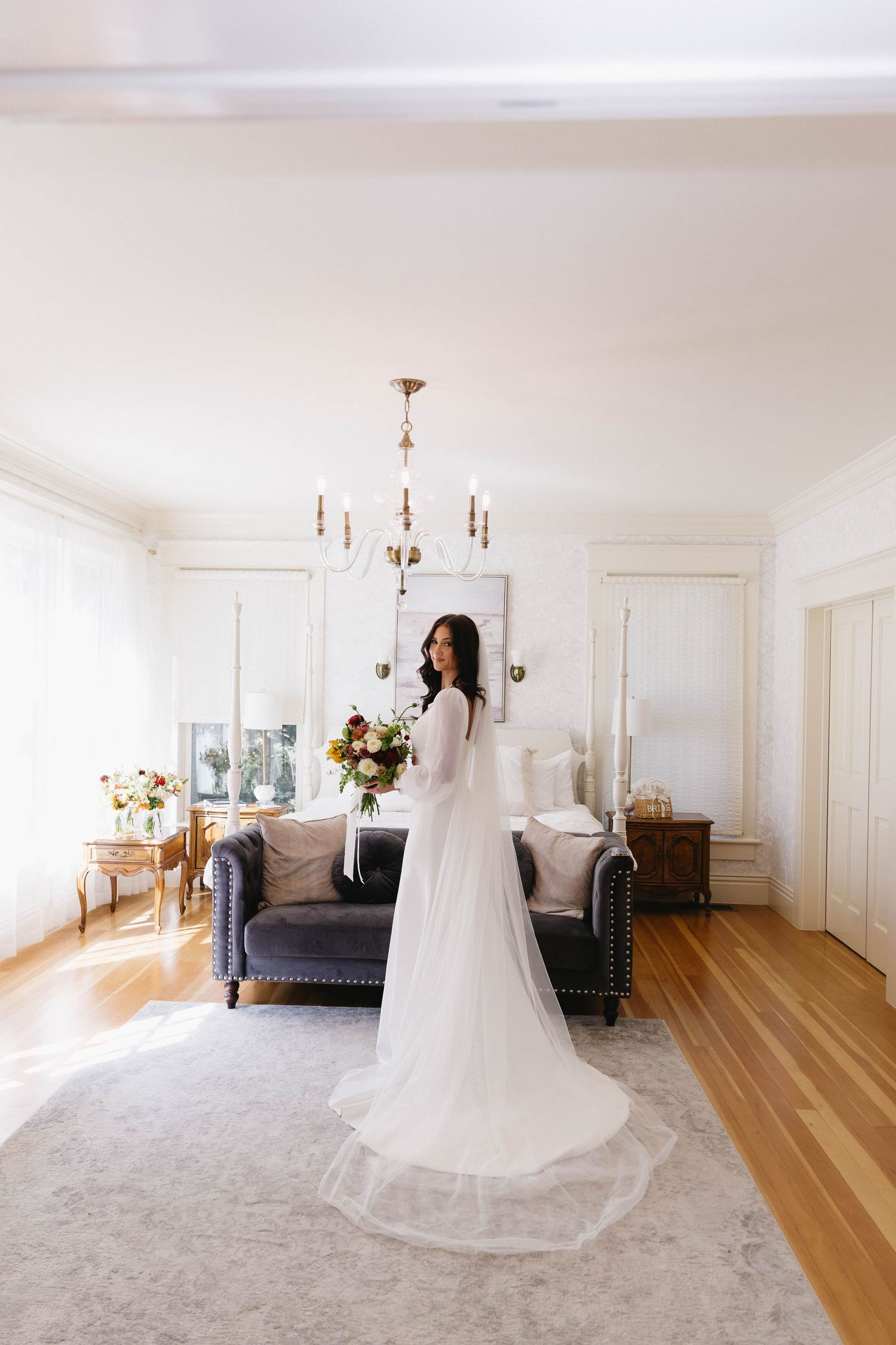 A flowing white wedding dress is displayed in an elegant room with hardwood floors and chandelier lighting.