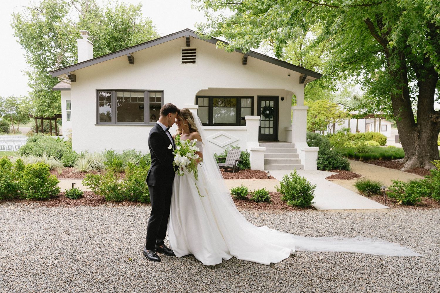 A wedding couple stands outside a white building surrounded by landscaped grounds.
