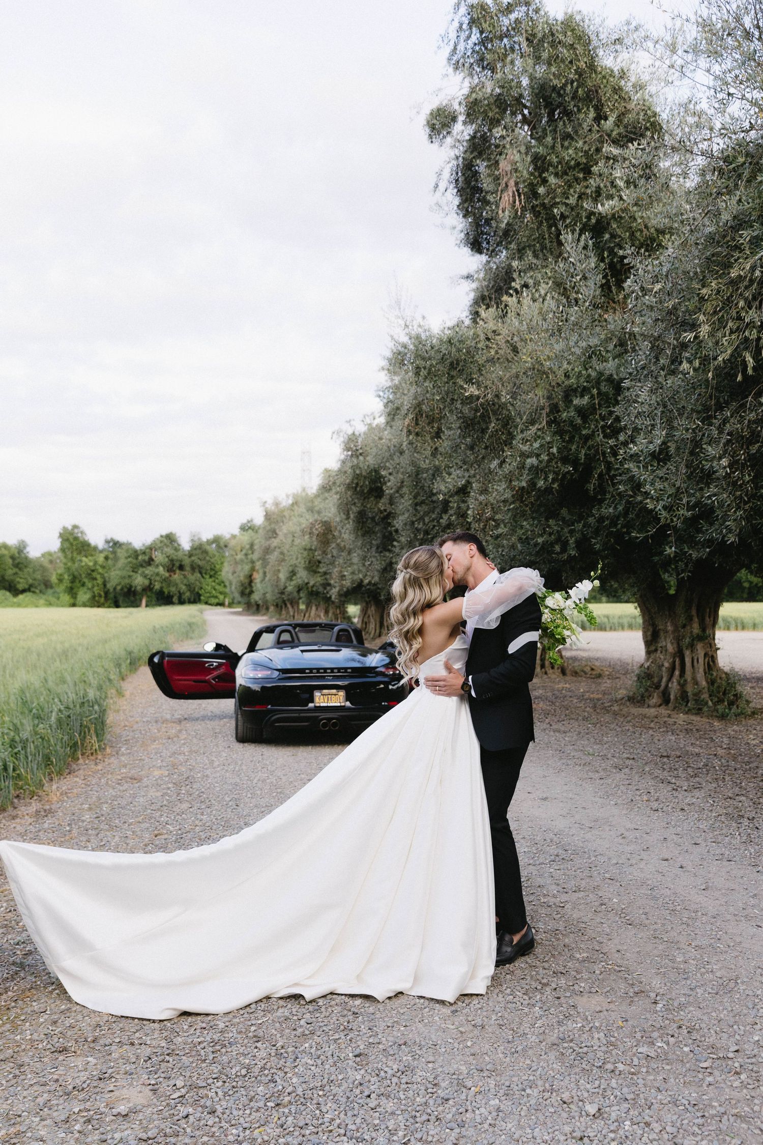 Wedding couple posing on a gravel path with olive trees and a black sports car in the background.