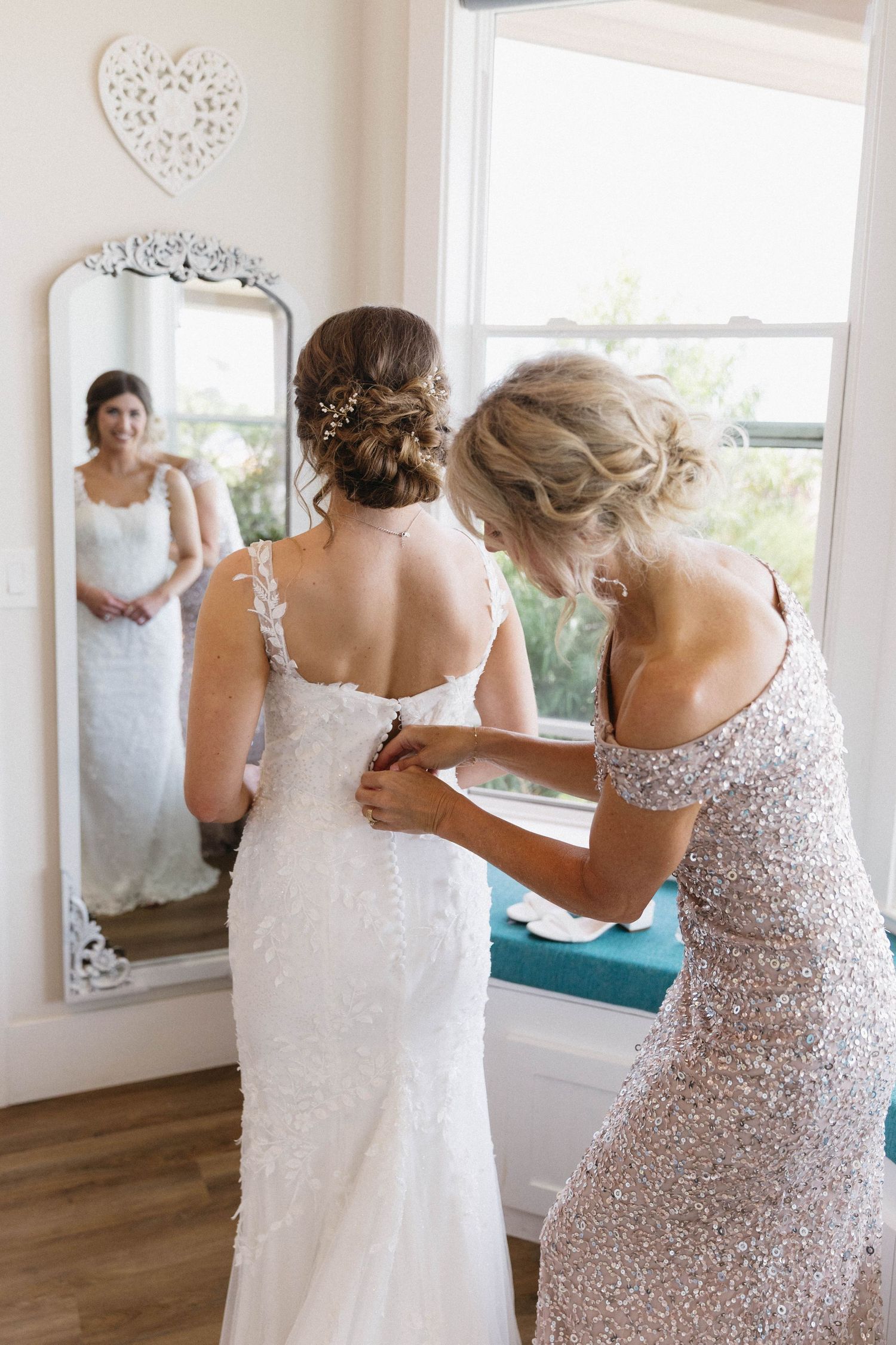 Wedding preparation scene showing dress fitting in a bright room with large windows.