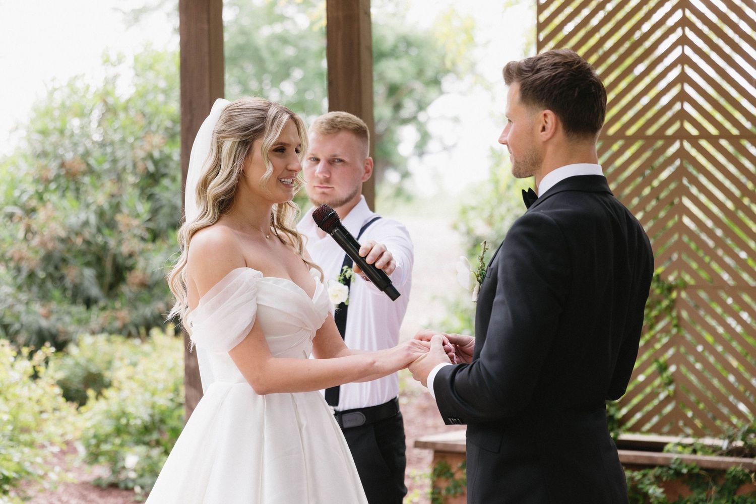 Wedding ceremony moment under wooden pergola with couple and officiant during vow exchange.