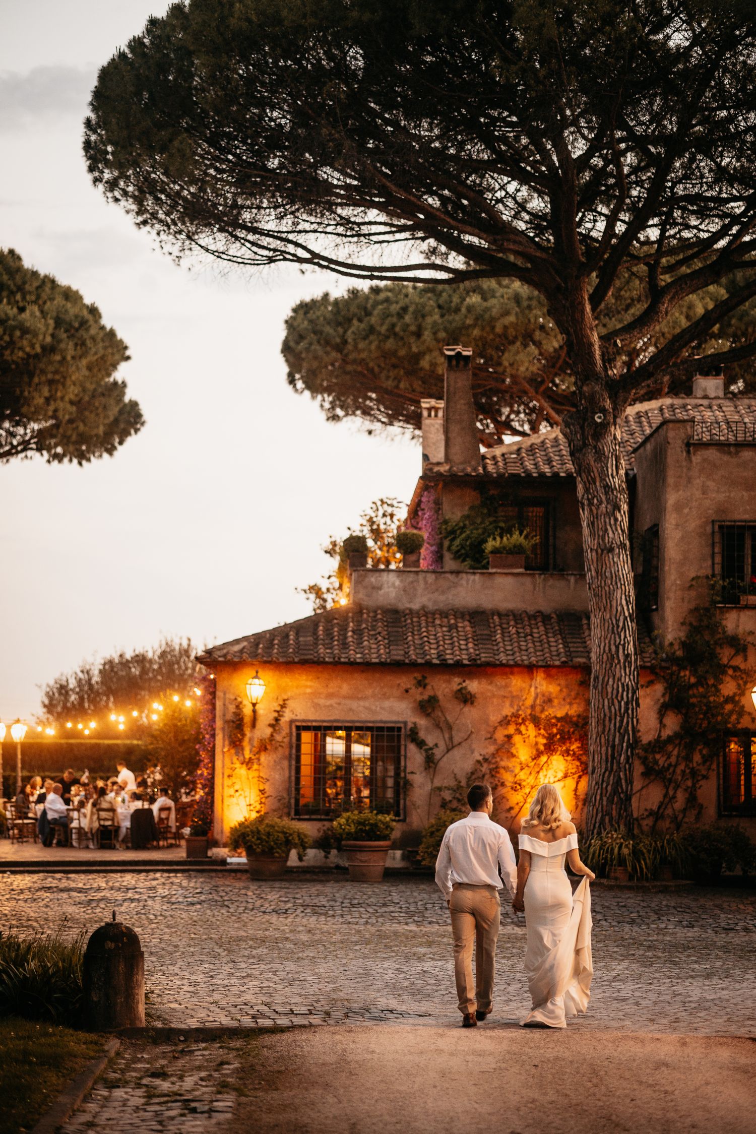 Couple walking toward rustic Italian villa with string lights at dusk.