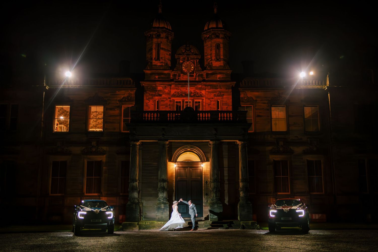 Dramatic nighttime wedding photos with car headlights illuminating a historic building exterior.