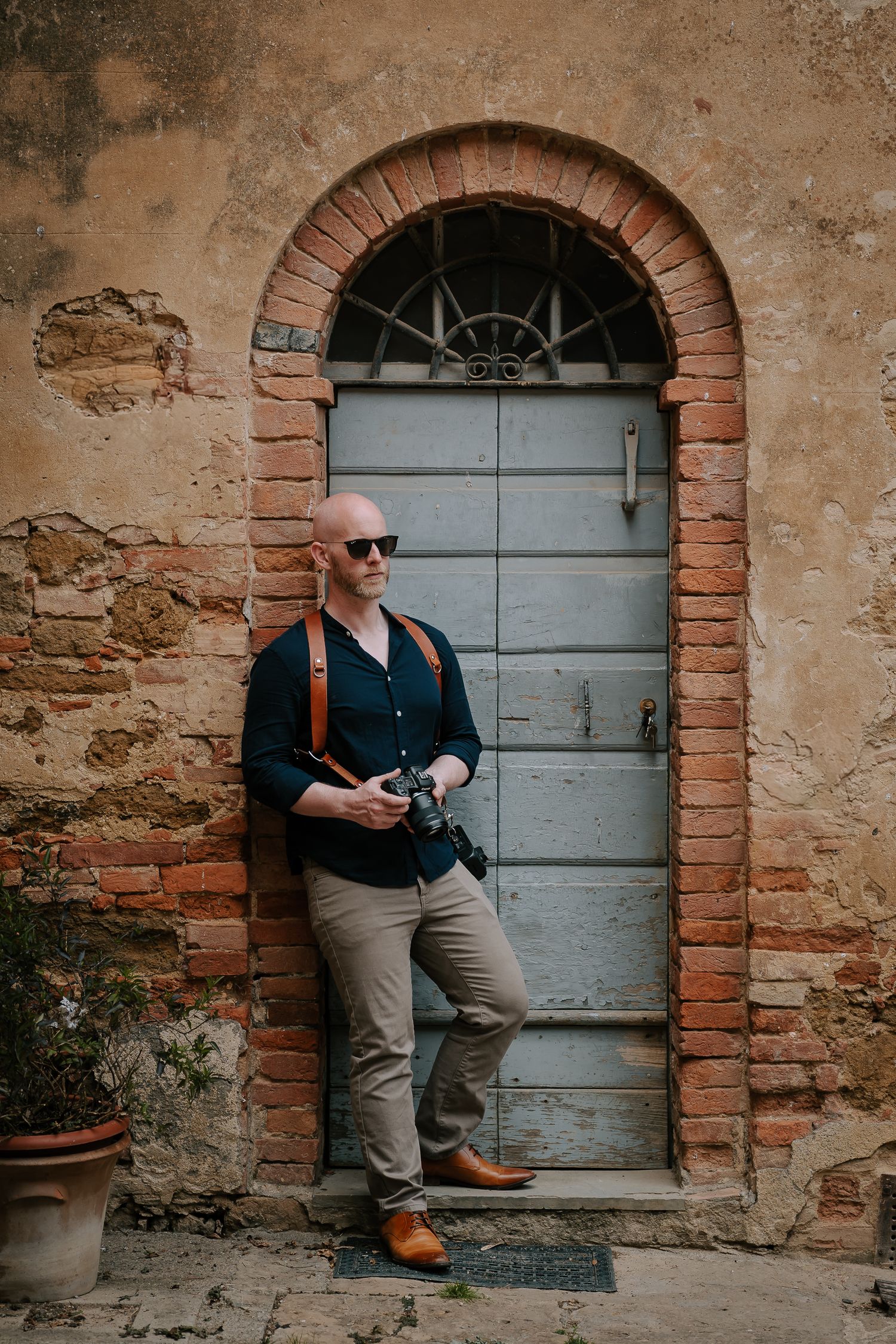 Photographer standing in rustic archway with vintage camera and casual professional attire.