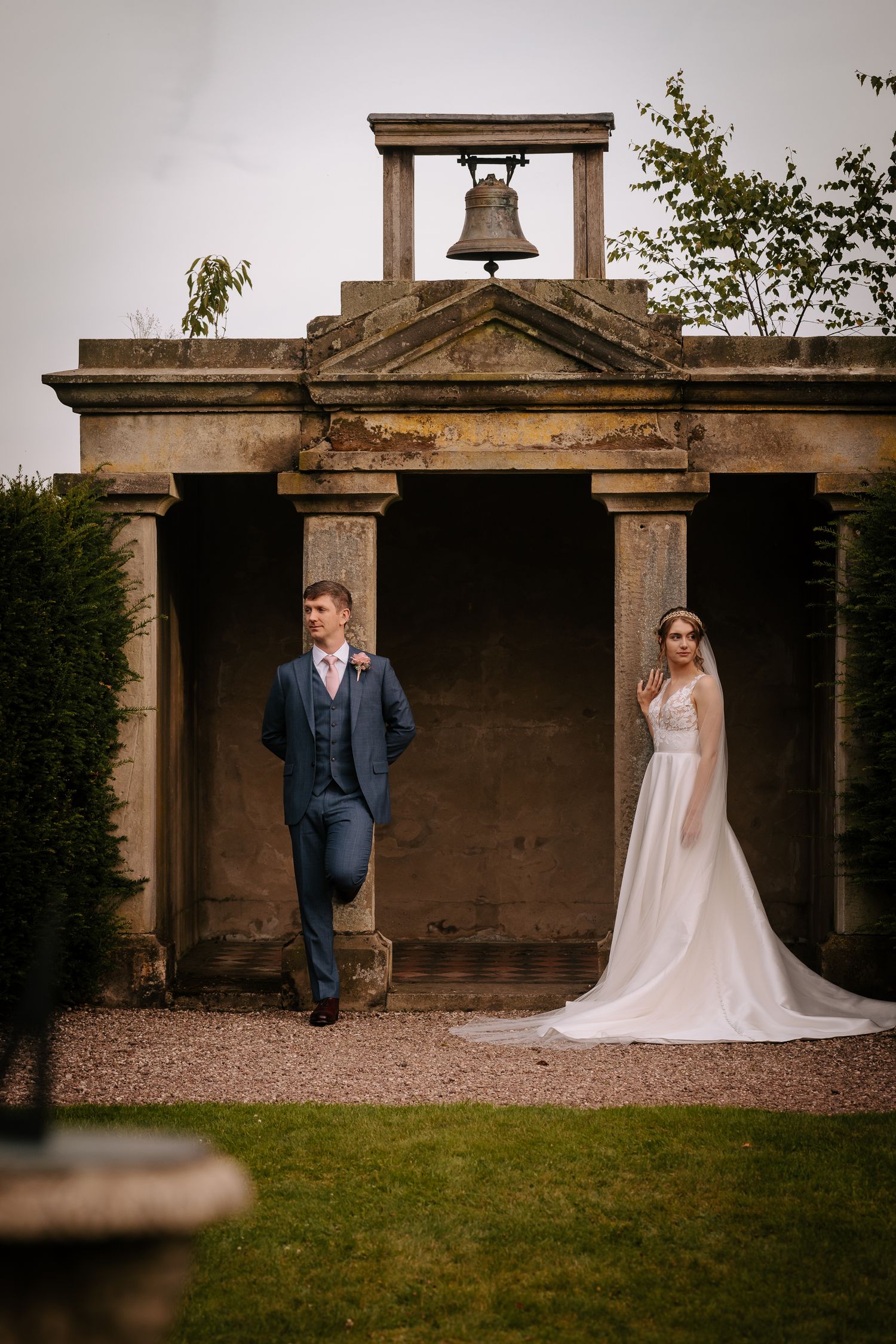 Couple in wedding attire pose separately near stone bell tower structure in garden setting.