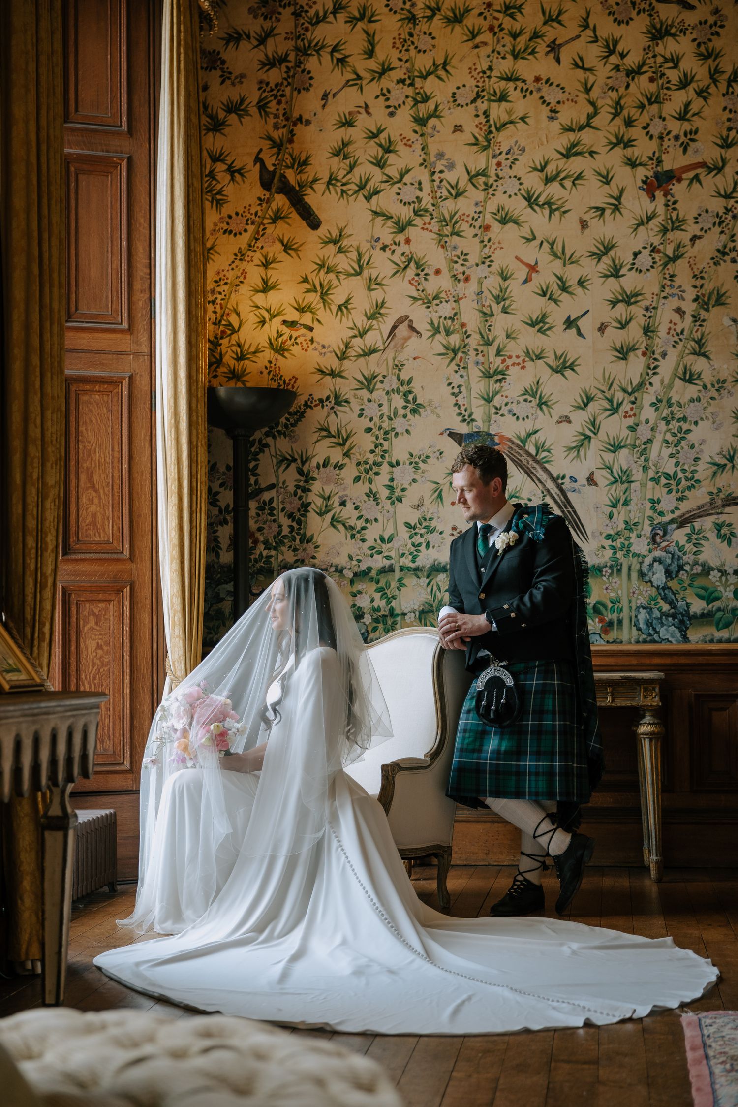Wedding photo in room with elegant floral wallpaper featuring traditional Scottish attire.