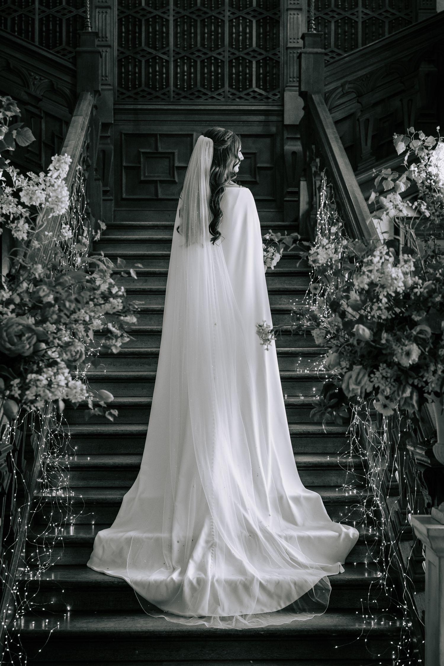 Dramatic black and white photo of bride in flowing dress on grand staircase with floral arrangements.