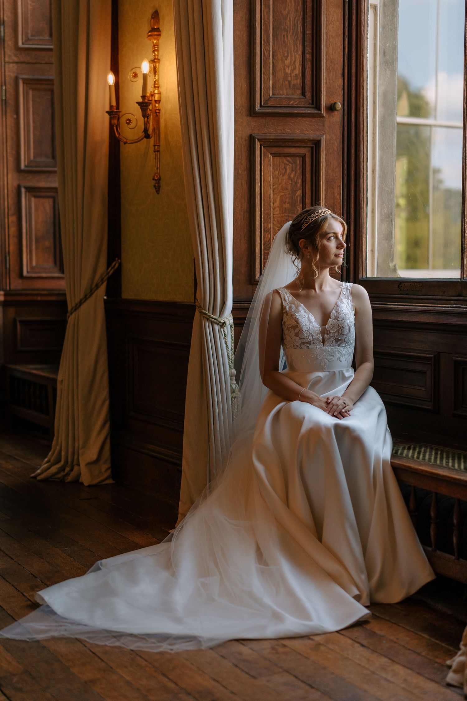 Elegant bride in lace wedding gown sits by window in historic room with ornate woodwork and wall sconce.
