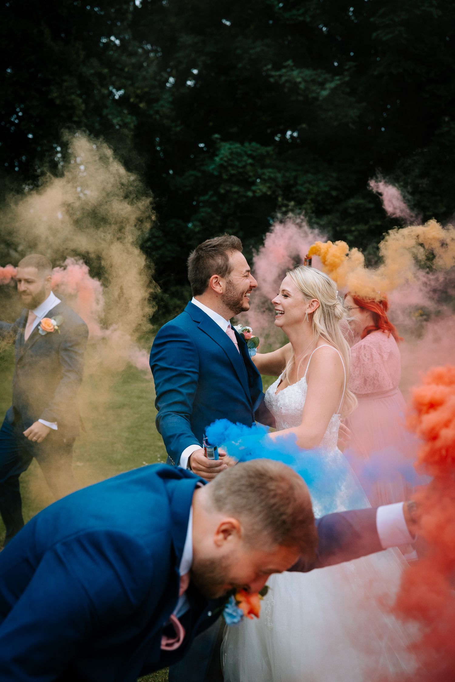 Wedding party celebrates with colorful smoke bombs creating vibrant clouds in outdoor setting.