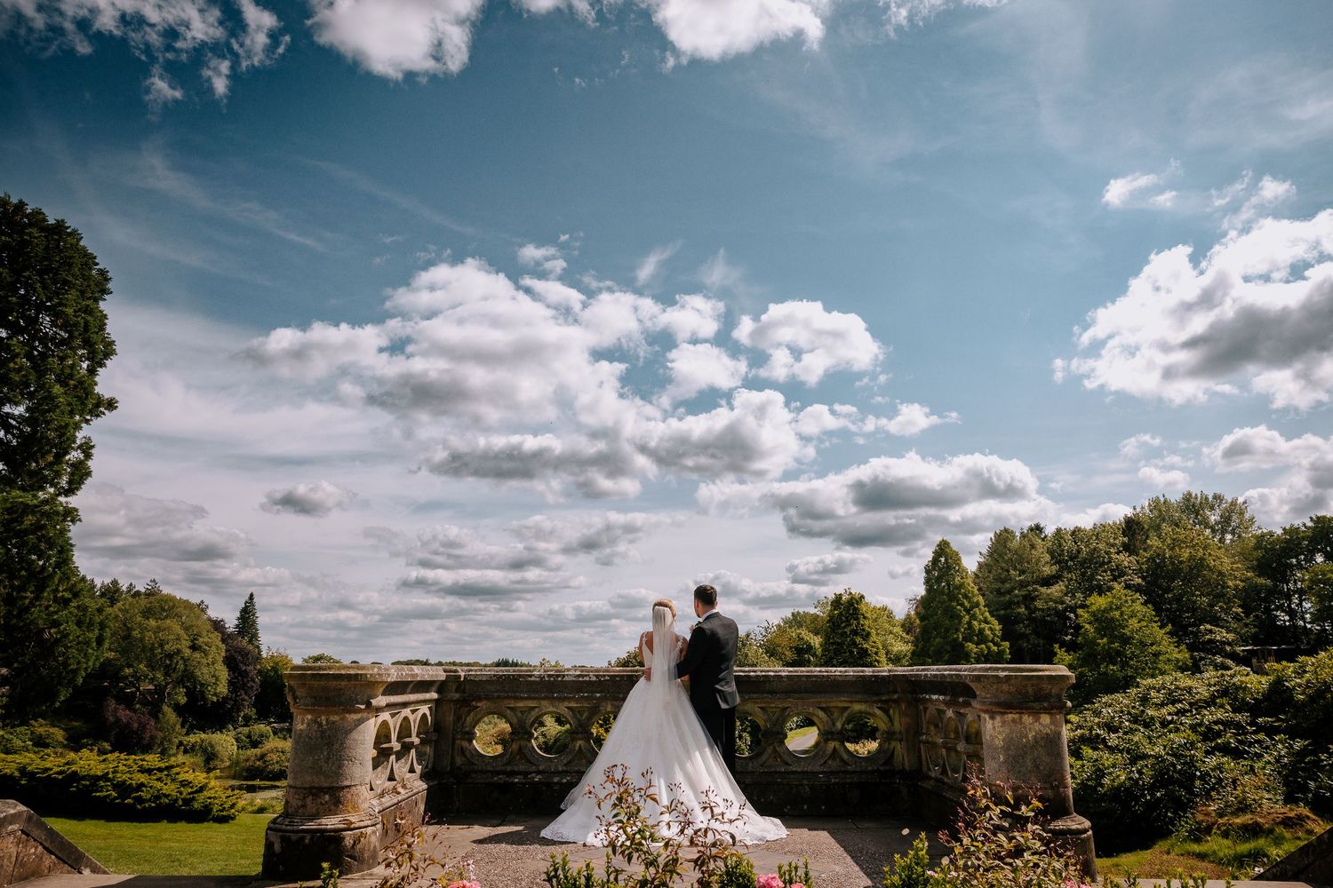 Wedding couple viewed from behind overlooking garden landscape on sunny day.
