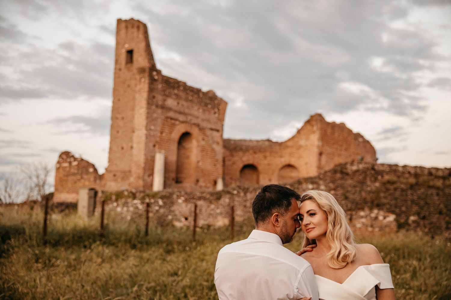 A couple embraces in front of ancient castle ruins during a warm sunset.