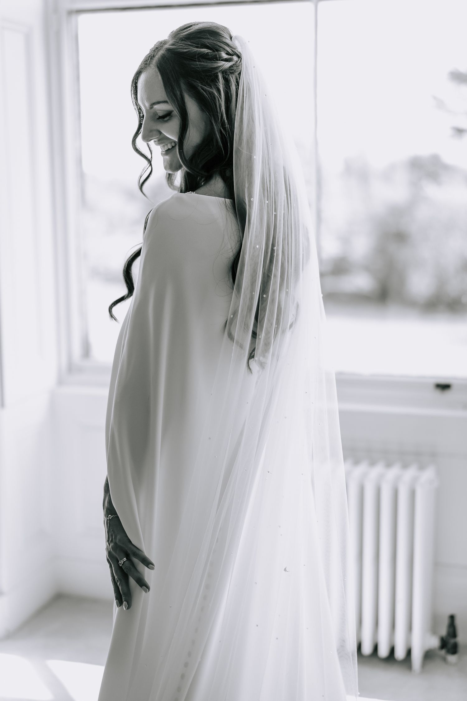 Ethereal black and white portrait of bride in flowing veil by window.