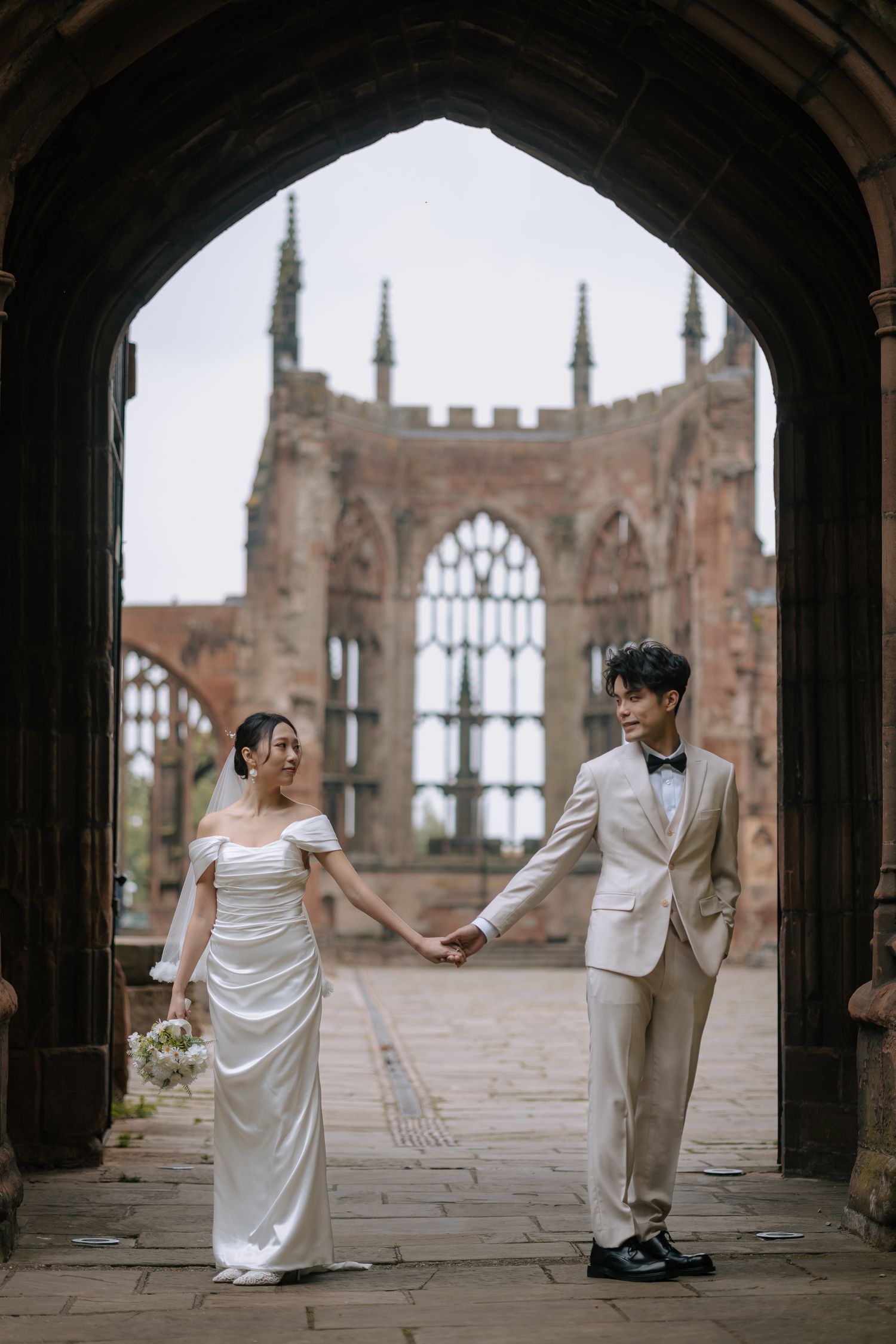 Couple in elegant wedding attire holding hands under a gothic cathedral archway.