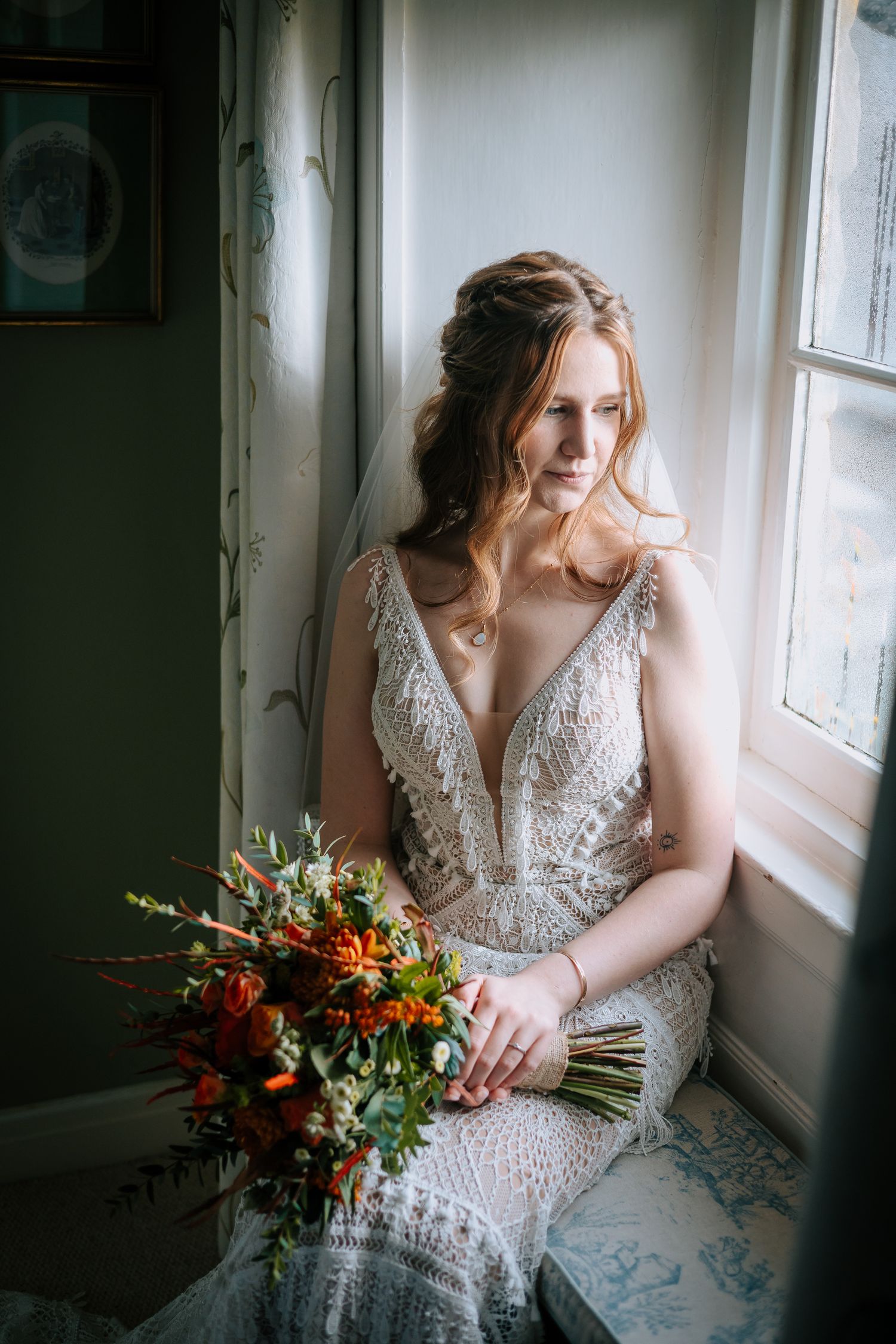 Bride in lace wedding dress poses by window with autumn-colored bouquet.