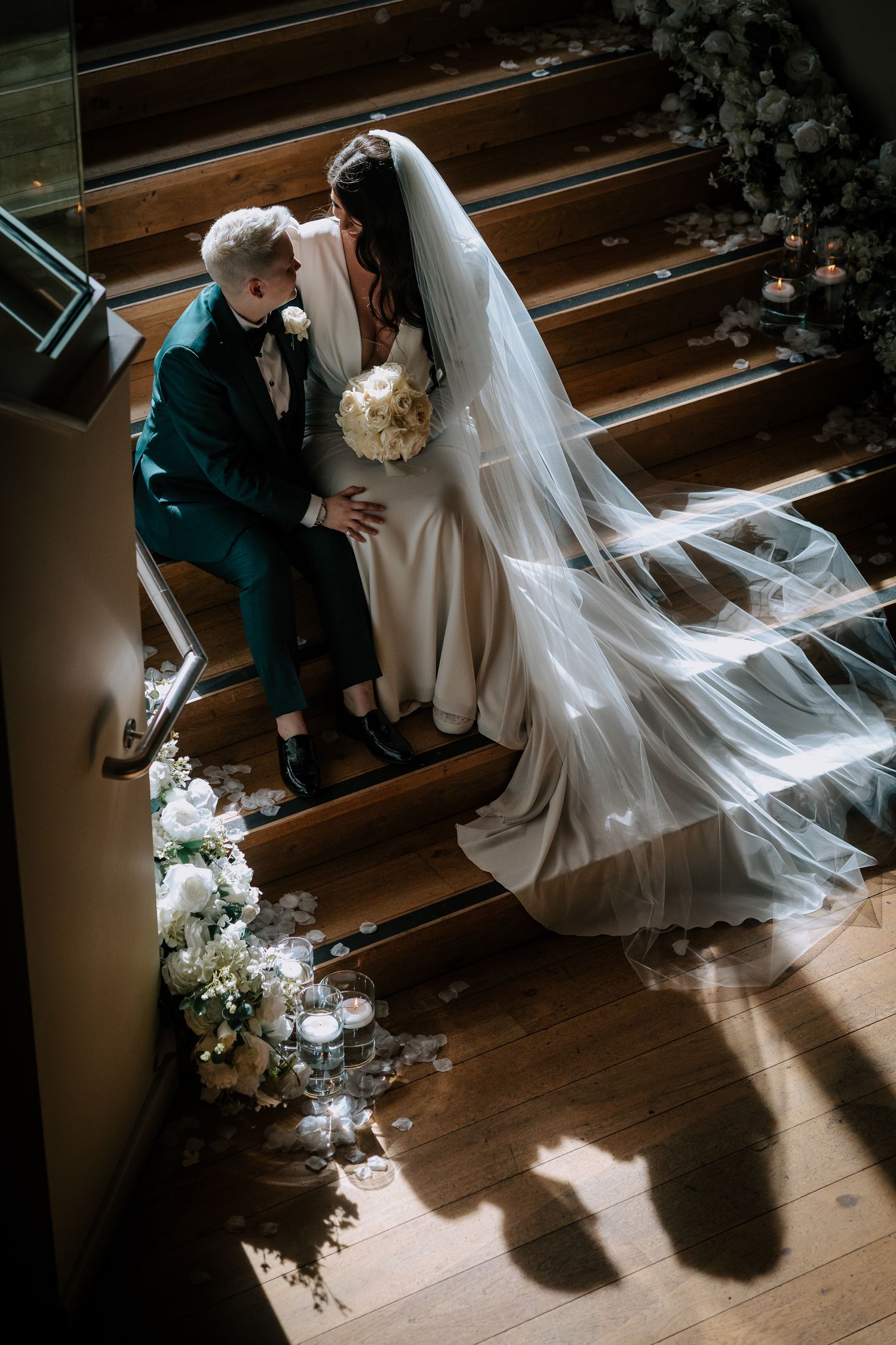 A dramatic wedding moment captured on wooden stairs with flowing veil and floral decorations.