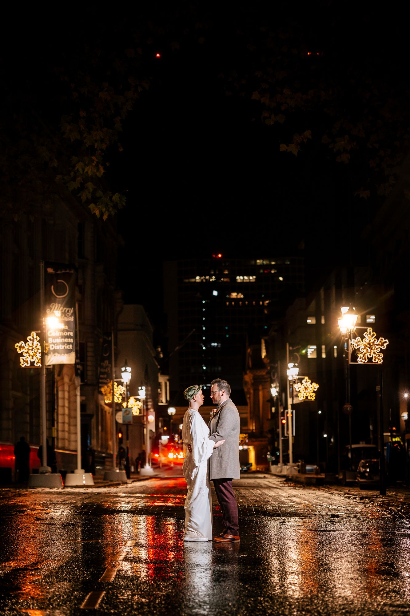 Romantic wedding photo on rainy city street with illuminated decorations.
